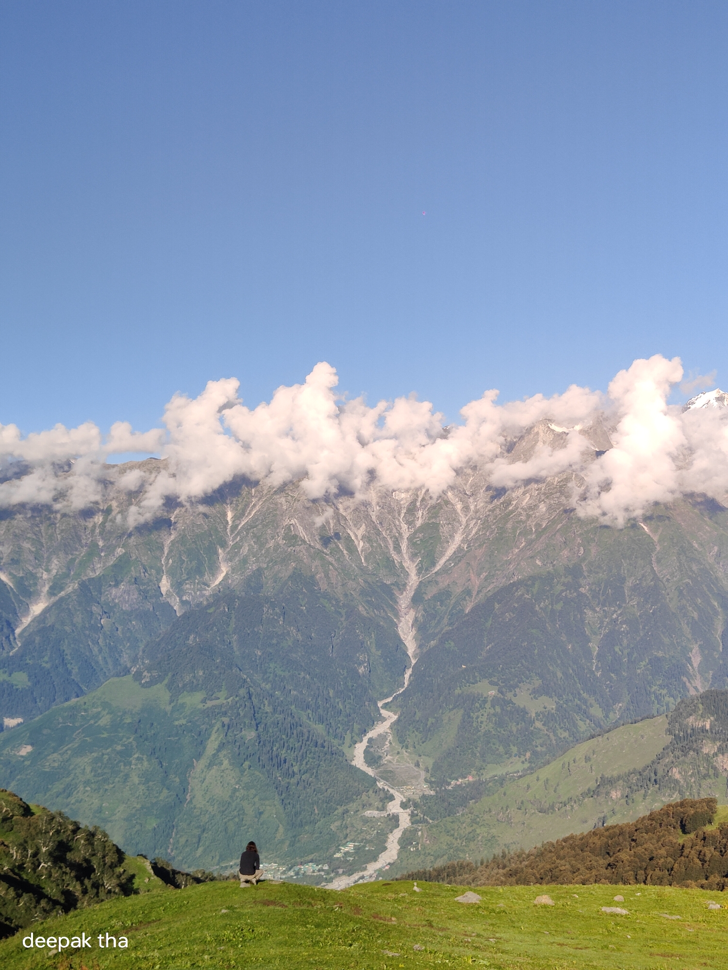 The image depicts a vast mountain landscape under a clear blue sky with some scattered clouds. In the foreground, a person is seated on a grassy knoll, facing away from the camera, gazing at the majestic mountains. The mountains themselves are imposing, with rugged slopes partially covered in green vegetation and some rocky outcrops. A river snakes its way through the valley below, adding a dynamic element to the scene. The overall impression is one of solitude and awe-inspiring natural beauty. The text "deepak tha" is visible in the bottom left corner of the image, likely indicating the photographer's name or a watermark.