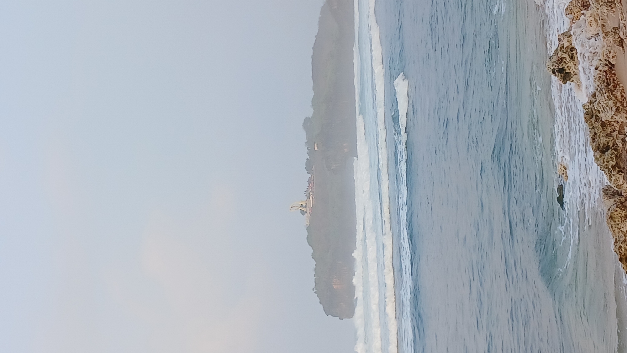 The image depicts a panoramic view of a coastal landscape, likely located in Tepus, Indonesia. In the foreground, a sandy beach is visible, adorned with scattered rocks and pebbles. The middle ground is dominated by the ocean, with gentle waves rolling towards the shore, creating white foam as they break. In the distance, a prominent cliff or headland juts out into the sea. The top of this landmass is covered with lush green vegetation. Perched on the highest point of the headland is a large, white, abstract statue or monument, likely a significant landmark. The sky above is overcast and hazy, suggesting a diffused light, possibly early morning or late afternoon. The overall atmosphere is serene and natural, emphasizing the rugged beauty of the coastline.