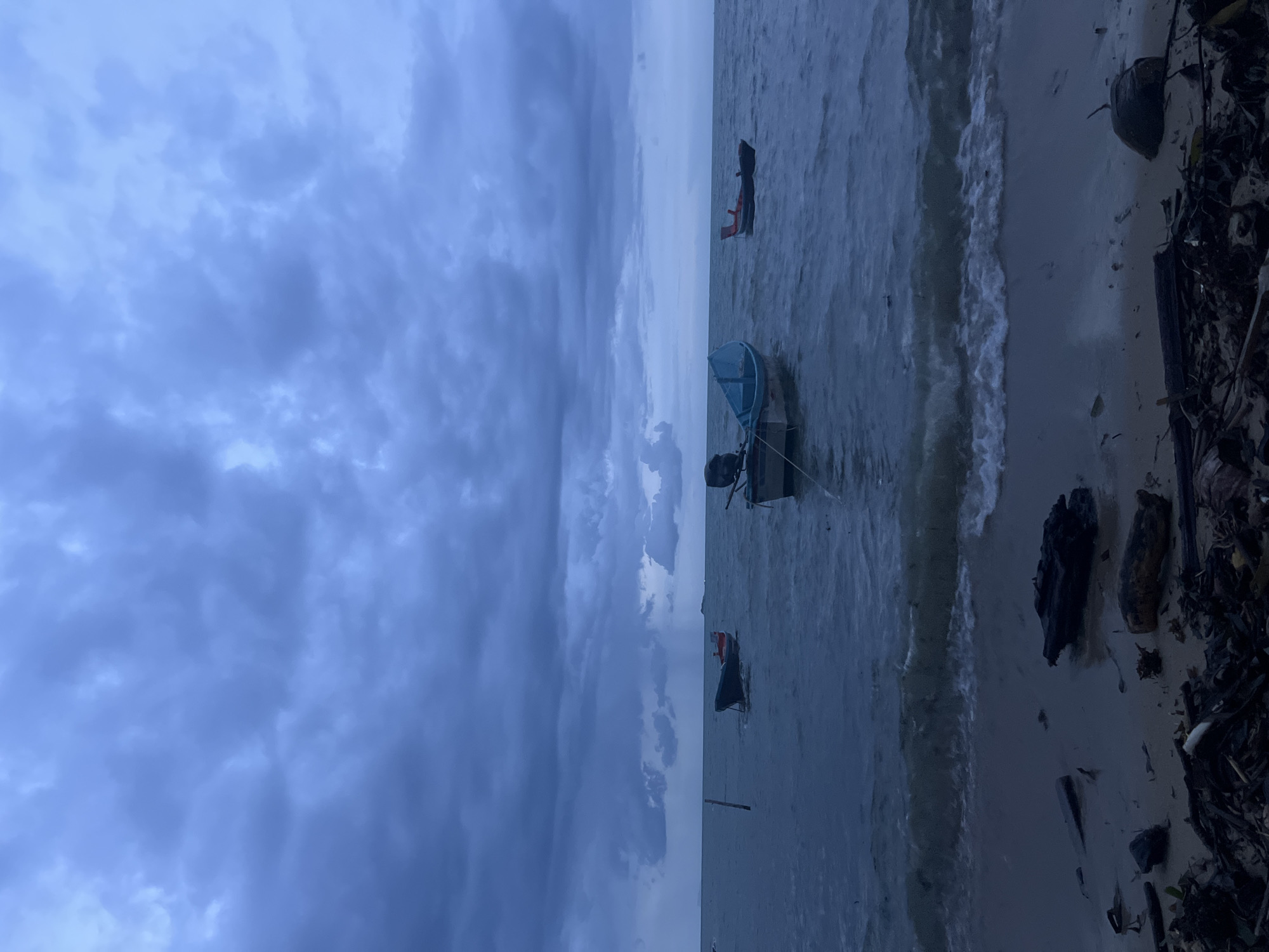 The image depicts a serene, yet somewhat ominous, seascape under a cloudy, twilight sky. The water is calm, with small waves gently lapping at a sandy shore littered with driftwood and debris. Three small boats are moored in the water. One is a dark hull with a red and white interior, closer to the shore and slightly to the left. Another, a light blue boat with a dark motor, is in the center of the frame, further out. A third, a dark boat with a red section, is visible to the right, also further out. The sky is dominated by heavy, dark clouds that suggest an approaching storm, with visible rain shafts in the distance. The overall mood is one of quiet solitude, tinged with the anticipation of inclement weather. The scene appears to be a coastal area, likely a beach, with no people visible. The lighting is dim, characteristic of dusk or an overcast day.