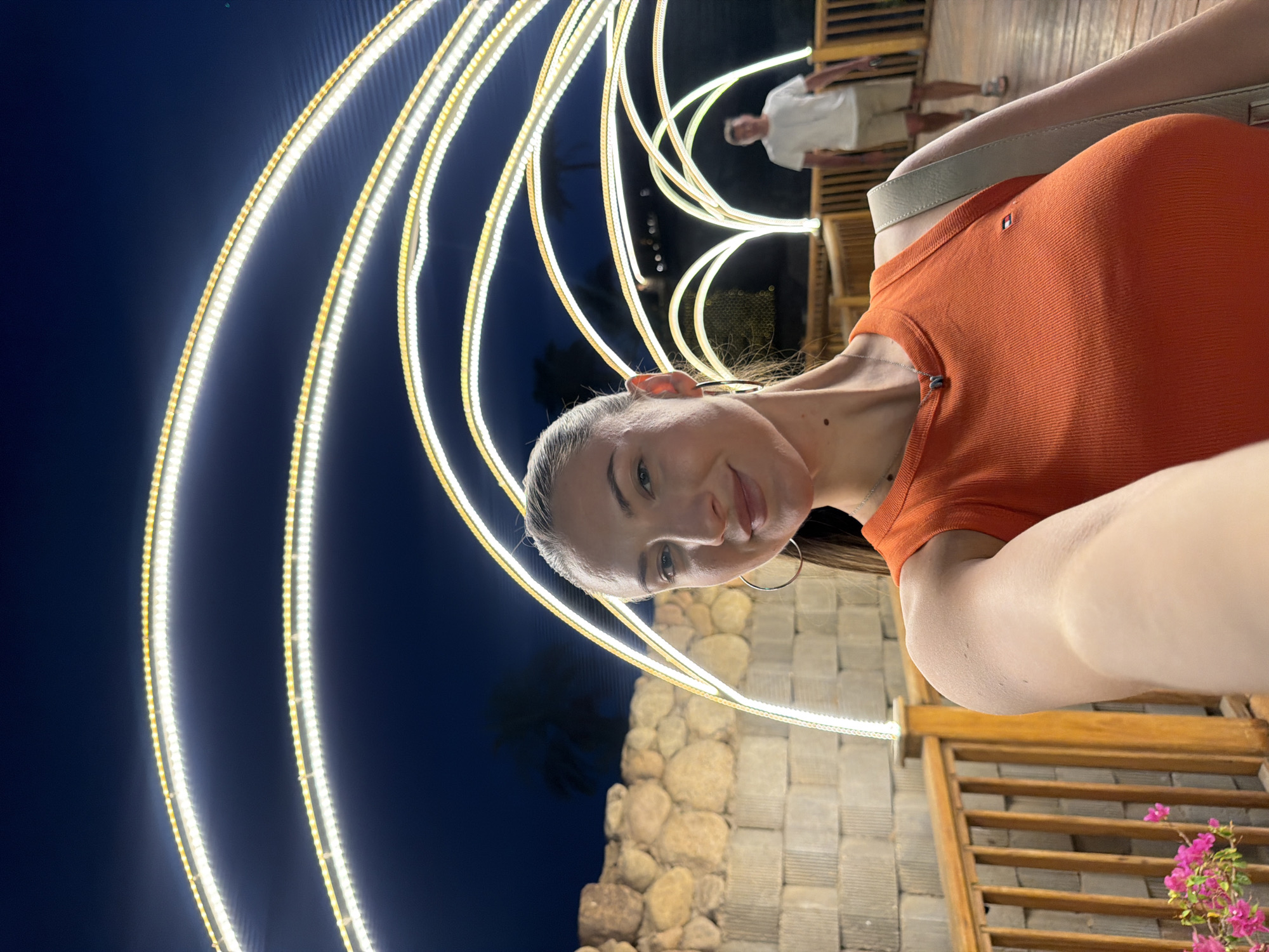 A woman takes a selfie at night in Sharm El Sheikh, Egypt, standing on a wooden boardwalk. She is wearing an orange tank top and large hoop earrings. Behind her, a man in a white t-shirt and shorts walks away. The scene is illuminated by decorative arched lights that extend into the darkness. A stone wall is visible to the left of the woman, and a wooden railing borders the boardwalk. Pink flowers are in the lower left corner of the frame.