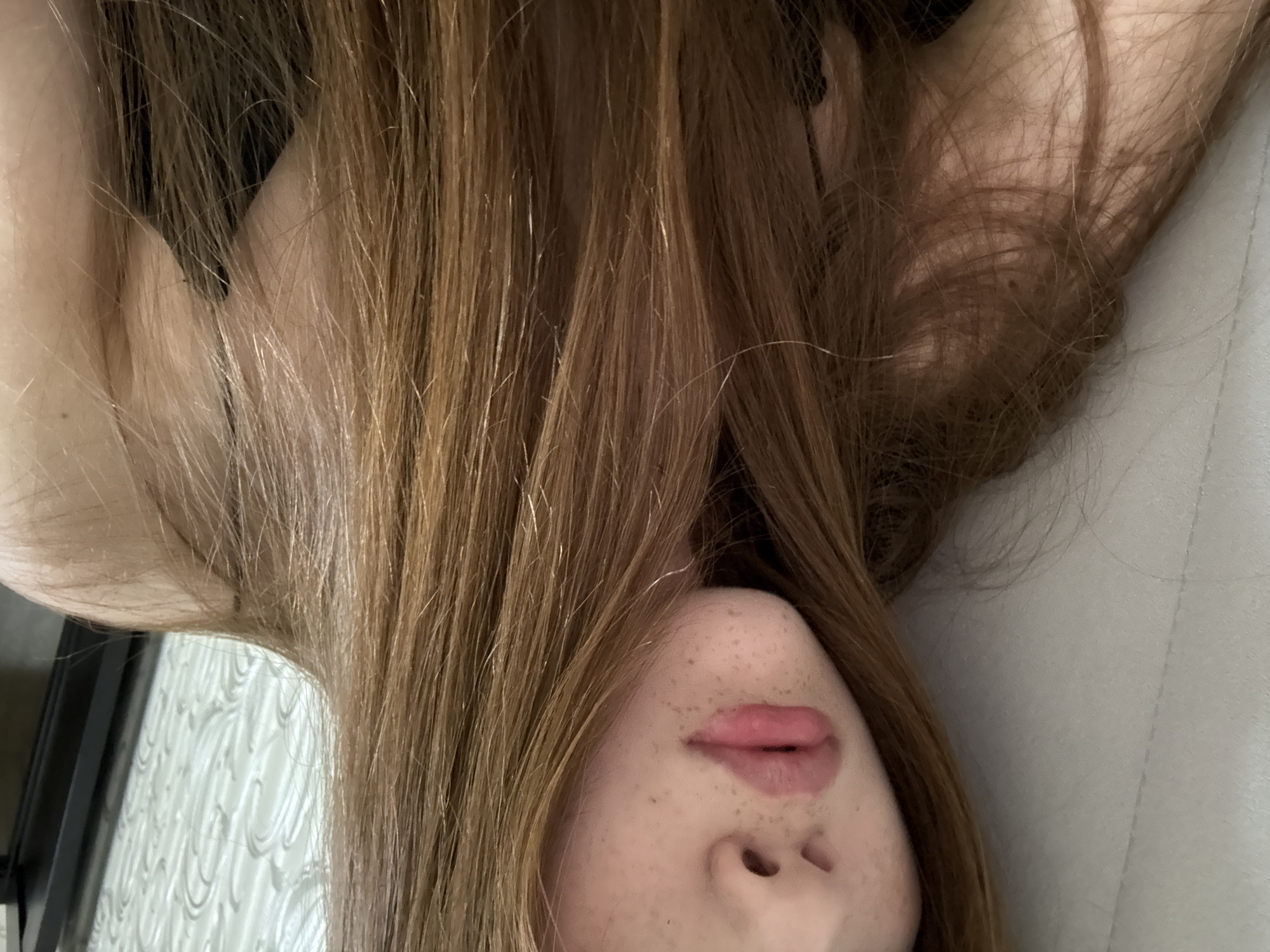 The image presents a close-up, rotated view of a person, likely female, lying down in an indoor setting in Naberezhnye Chelny, Russia. The central focus is on the person's long, light to medium brown hair, which cascades around and partially over their face. The lower portion of the face is visible, showing the nose, lips, and chin, all marked by numerous freckles. The lips are a natural pinkish-red color.
To the left of the person's head, a light-colored surface with a distinct wavy pattern is visible, possibly a curtain or textured wall, alongside a dark vertical structure that could be a window or mirror frame. On the right side of the frame, a soft, light grey material, likely a pillow or bedsheet, fills the background. The lighting is soft and indirect, suggesting a relaxed atmosphere. No text is visible in the image. The composition suggests a personal, candid moment of rest or contemplation.