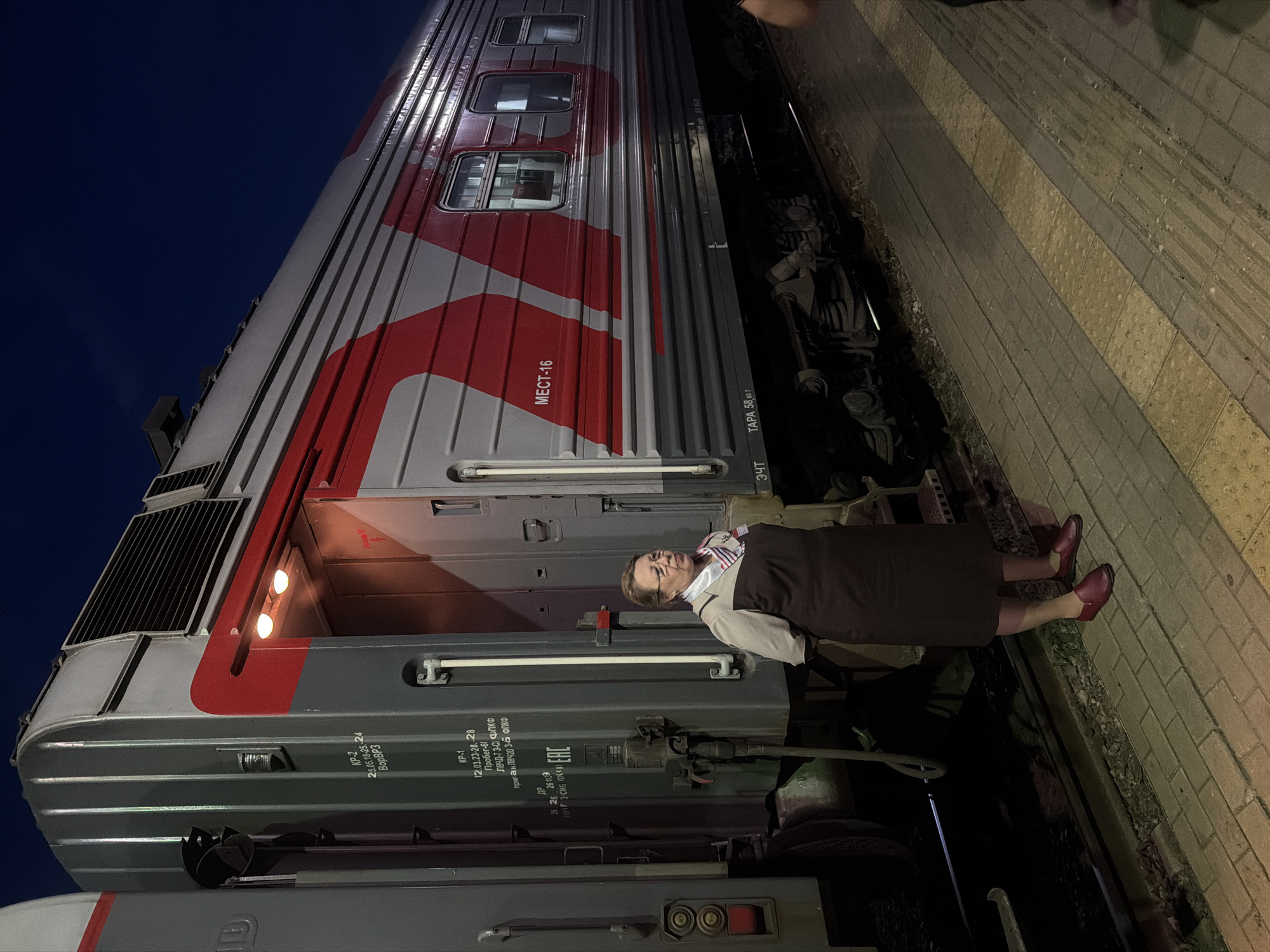 A woman stands on a train platform at the Balashov-1 train station in Balashov, Russia. The woman is wearing a dark brown uniform dress, a light-colored collared shirt, and a scarf. She has glasses on and her hair is pulled back. She is standing next to a train car that is gray with red markings. The train car has several windows and appears to be an older model. The platform is paved with bricks. The time of day is dark, suggesting it is nighttime or very early morning. There are lights on the train car, illuminating the area. The environment is an outdoor train station with tracks visible next to the platform.