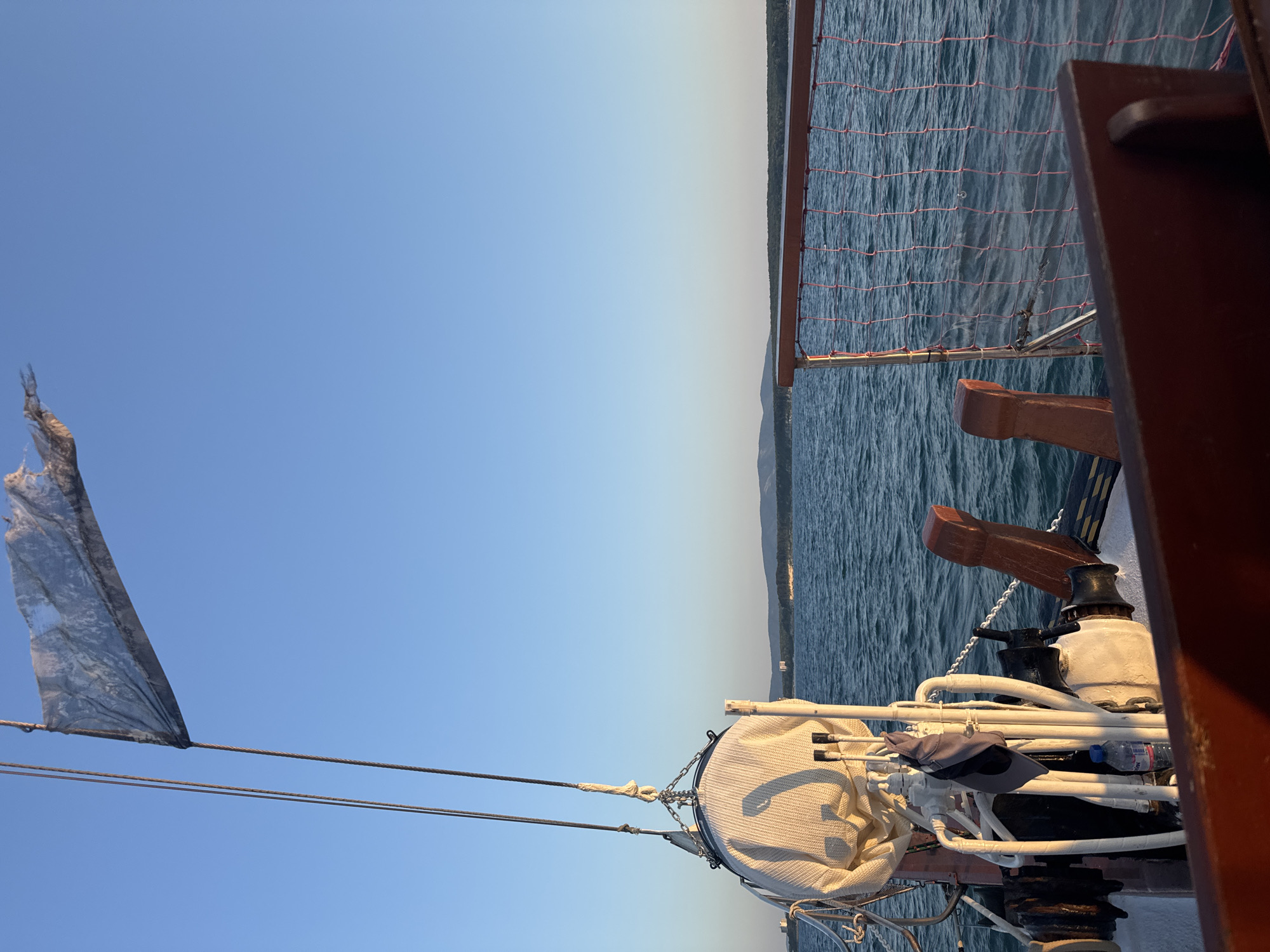 The image shows a view from the deck of a sailboat at sea, looking towards the horizon. In the foreground, parts of the boat's rigging and deck are visible, including a sail cover, ropes, and wooden railings. A tattered flag flutters from a mast against a clear blue sky. In the distance, a low-lying coastline with hills and trees can be seen, suggesting the location is near land. The sea is calm with gentle waves. The scene appears to be set during sunset or sunrise, with the warm light casting shadows on the boat and illuminating the water. The distant coastline appears to be part of the area around Primorsko, Bulgaria, a Black Sea resort town known for its beaches and natural beauty.