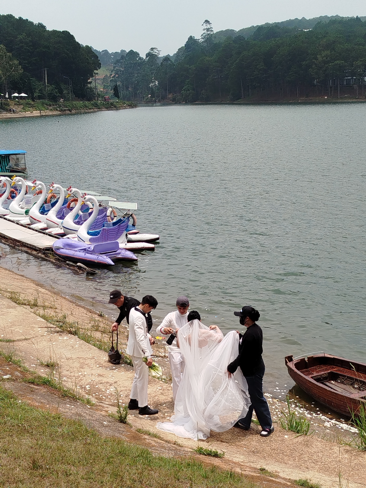 The image depicts a wedding photoshoot in Da Lat, Vietnam. A bride in a white gown and veil stands near the edge of Tuyen Lam Lake, attended by a groom in a white suit and three assistants. One assistant adjusts the bride's veil, another carries equipment, and the third appears to be holding the gown. A row of swan-shaped paddle boats is docked in the background. A wooden boat is partially visible in the water near the wedding party. The background features a lake surrounded by forested hills.