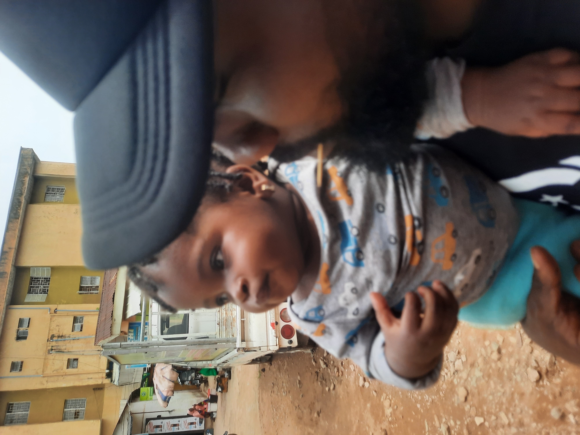 The image captures a man holding a baby outdoors in Jos, Nigeria. The man is wearing a dark cap, and his face is partially visible, suggesting he is looking down at the child. The baby is wearing a patterned gray shirt and blue pants. In the background, there are buildings with windows and shops, indicating an urban setting. The ground appears to be dry and dusty.