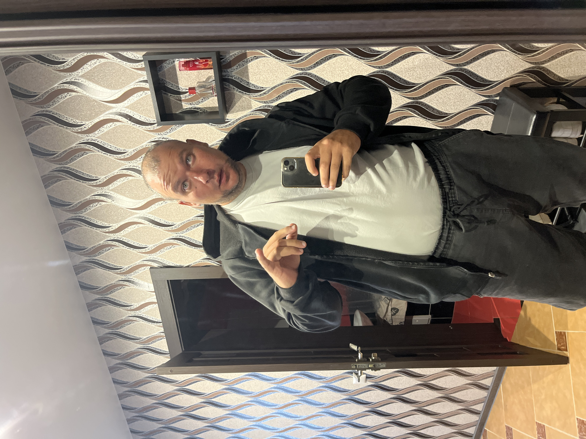 In the city of Minsk, Belarus, a bald man with light blue eyes stands in a room with patterned wallpaper. He is wearing a black hoodie over a light-colored t-shirt and dark sweatpants. He is holding a smartphone in his right hand, taking a mirror selfie. In his left hand, he gestures forward. To his left, a doorway is partially open, revealing a glimpse of what appears to be a closet or storage area with red bins. To his right, a small black shelf is mounted on the wall, holding two small bottles, one red and one clear, and what appears to be a spray bottle with a red label. The floor is tiled in a warm, golden hue.