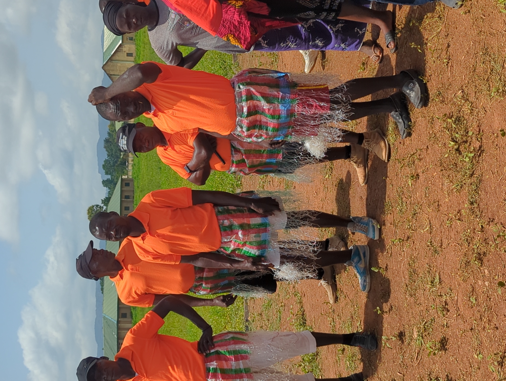 The image depicts a group of at least five men standing outdoors, likely in the city of Maigizo, Nigeria. All of the men are wearing bright orange shirts and knee-length skirts that appear to be made of a striped material and adorned with strands of white fringe. Their footwear varies, including black dress shoes, tan boots, and blue sneakers. Some of the men wear hats or caps. The background features a green field, buildings with green roofs, and hills in the distance beneath a partly cloudy sky. The ground appears to be a reddish-brown soil. The men are arranged in a line, with some of them making gestures like touching their heads or crossing their arms. The scene seems to capture a cultural performance or gathering of some kind.