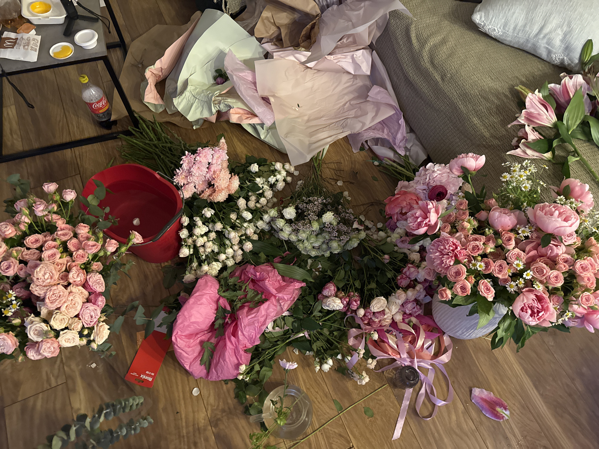 The scene depicts a wooden floor scattered with flowers and floral arrangements. The flowers are predominantly pink and white, including roses, peonies, and lilies. Some of the flowers are in a red bucket filled with water, while others are arranged in a gray vase. There is also a clear glass vase with a single flower in it. Floral wrapping paper in shades of pink, green, brown and white is strewn around the flowers, creating a chaotic and informal scene. Other objects include a partially consumed McDonald's meal on a nearby table with a Coca-Cola bottle, and a portion of a grey sofa visible in the upper right corner. A red paint sample card is near the bottom left. The location appears to be indoors, perhaps in a home. No people are visible.