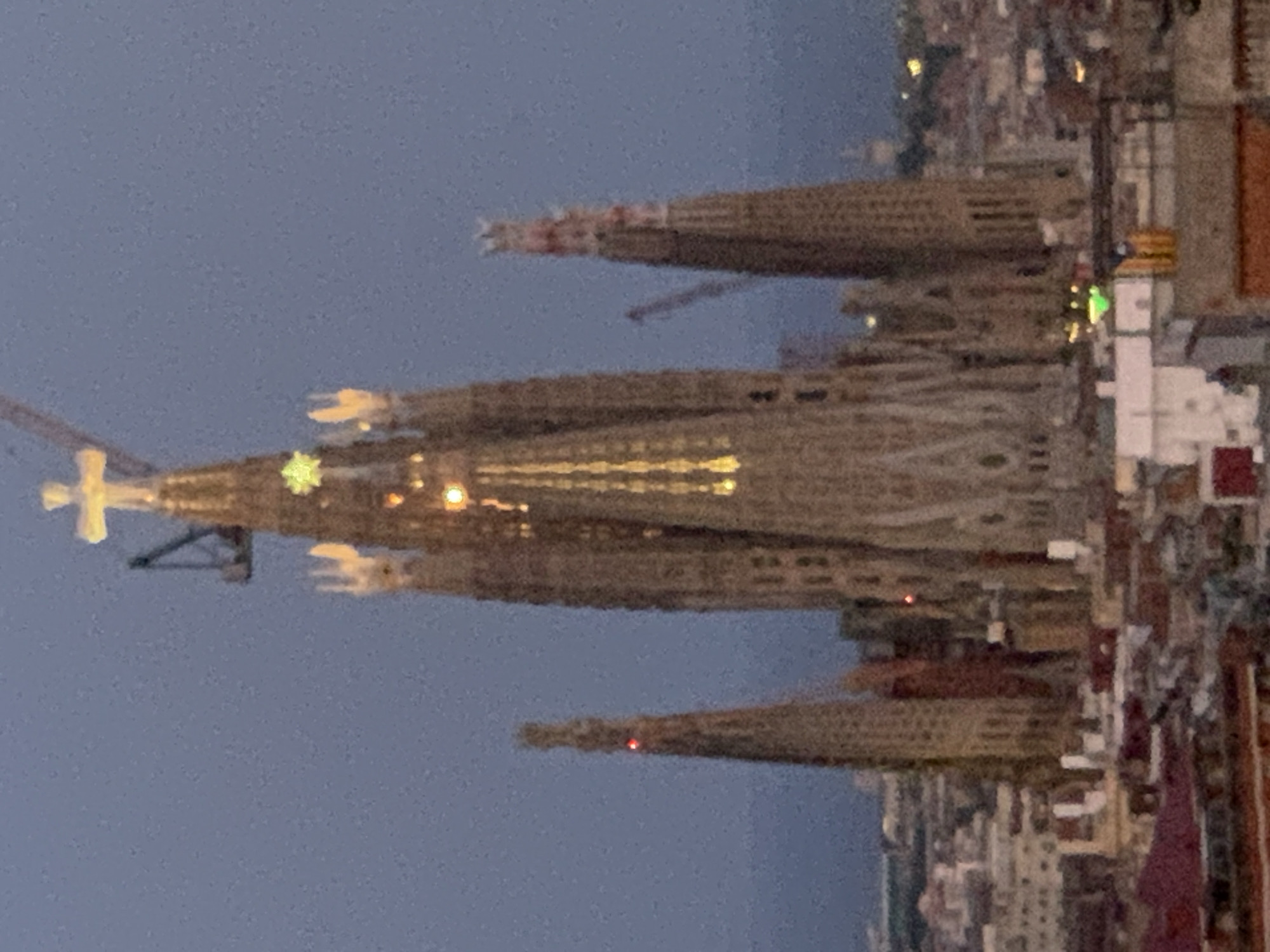 The image shows a view of the Sagrada Familia basilica in Barcelona, Spain, at dusk. The basilica's unfinished spires are visible, with construction cranes present. The sky is a deep blue, and lights are beginning to illuminate the building and the surrounding city. The foreground features rooftops of residential buildings.