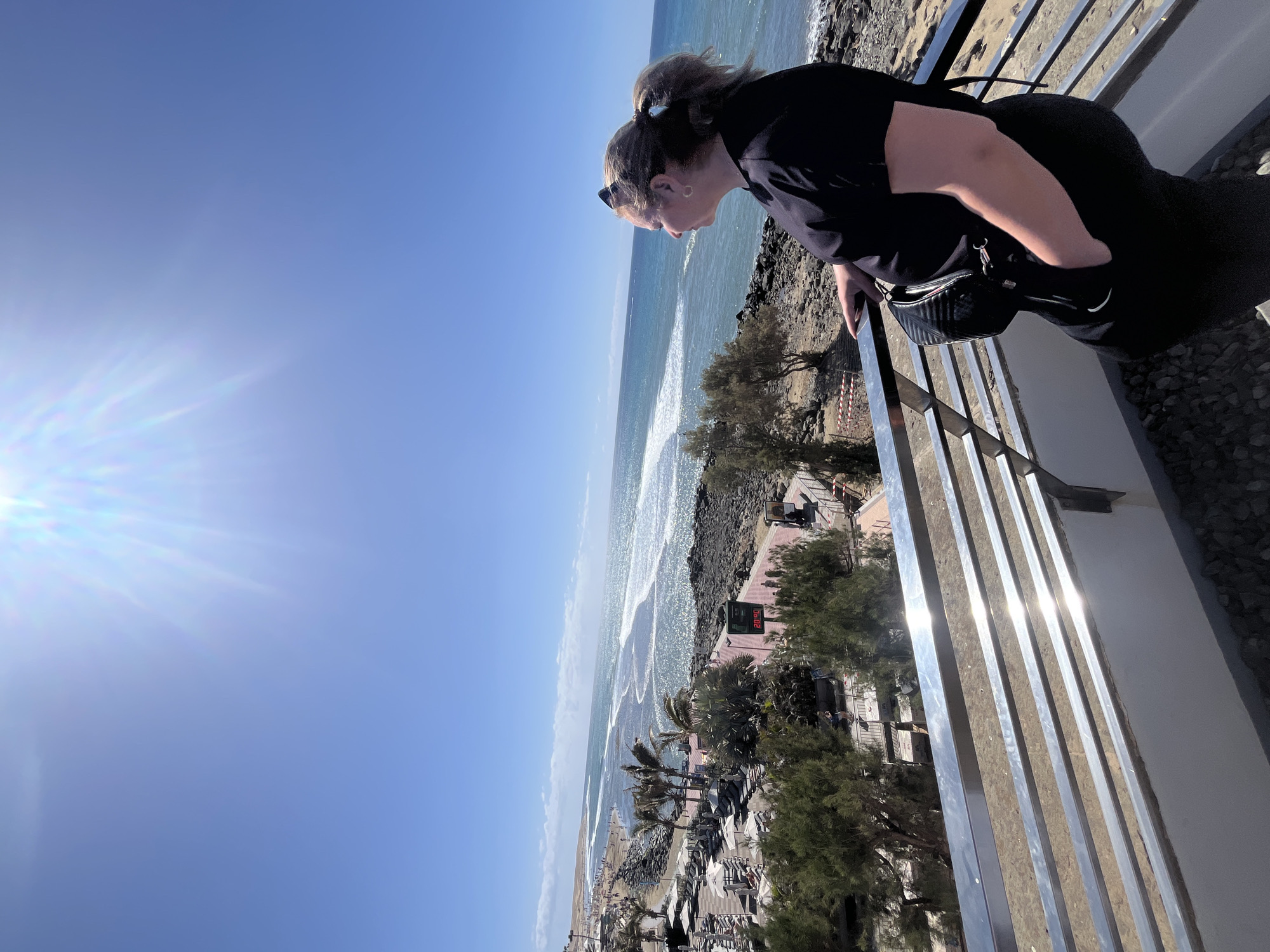 A high-angle, slightly rotated outdoor shot shows a woman in black clothing leaning over a railing, looking down at a beach scene. The location is likely Maspalomas, Spain, as indicated by the beach and ocean landscape. The woman has her hair up in a ponytail and is wearing sunglasses. She has one hand in the pocket of a small black crossbody bag. The railing she's leaning on is made of vertical metal bars. Below, the beach has palm trees, sunbathers, and people walking. The sky is bright blue with the sun shining. The ocean has visible waves.