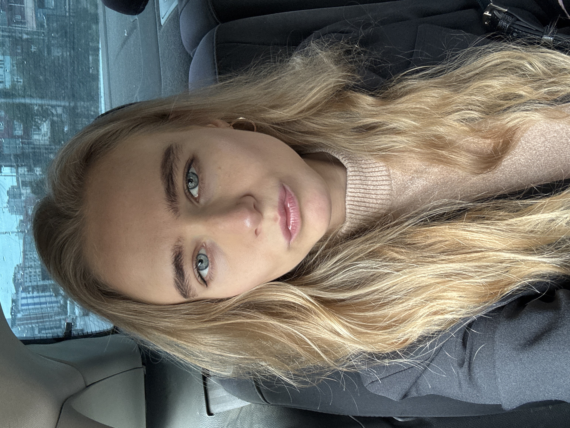 A young woman with long blonde hair is seated in a car. The car appears to be in an urban setting, given the buildings visible through the car window. The woman is wearing a light pink sweater and a dark jacket. She is looking at the camera with light blue eyes.