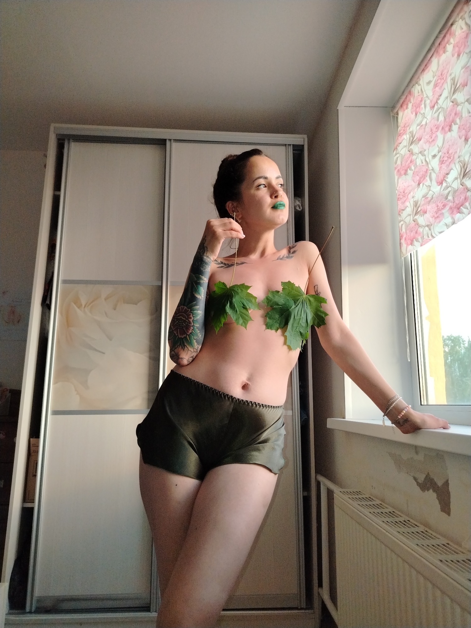 A woman is depicted indoors, standing in a room and leaning against a window sill, looking contemplatively out the window. She has dark hair pulled back and wears striking green lipstick. Her upper body is covered only by two large green maple leaves, while she wears dark olive green shorts. Her left arm is extensively covered in tattoos, featuring floral and leafy designs with a prominent red flower on her forearm. She also has smaller tattoo elements on her upper chest and shoulder. Her right arm, adorned with several bracelets including a strand of pink beads, rests on the window sill.
In the background, to her left, is a large, light-colored sliding door wardrobe with panels, some exhibiting a subtle patterned design. To her right, a window is partially covered by a roller blind with a pink floral pattern. Beyond the window, hints of green trees and a building with a yellow facade are visible under natural daylight. Below the window, a white radiator is present, and the wall above it shows patches of peeled paint or wallpaper. The scene provides a glimpse into a private indoor setting in Noviy, Russia. No visible text, other than the tattoos, is discernible.