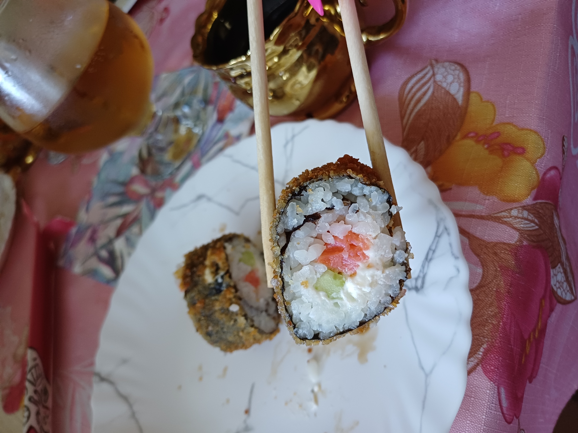 A close-up, eye-level, outdoor shot captures two pieces of deep-fried sushi rolls on a white plate with a marble pattern, placed on a pink tablecloth with a floral design. One roll is held by wooden chopsticks, revealing its filling of rice, cream cheese, cucumber, and salmon. Another sushi roll rests behind it on the plate. In the background, a glass of amber-colored liquid, possibly beer or tea, sits next to a golden ornamental cup. The lighting is bright, casting soft shadows. The scene is set on a table, likely for a meal.
