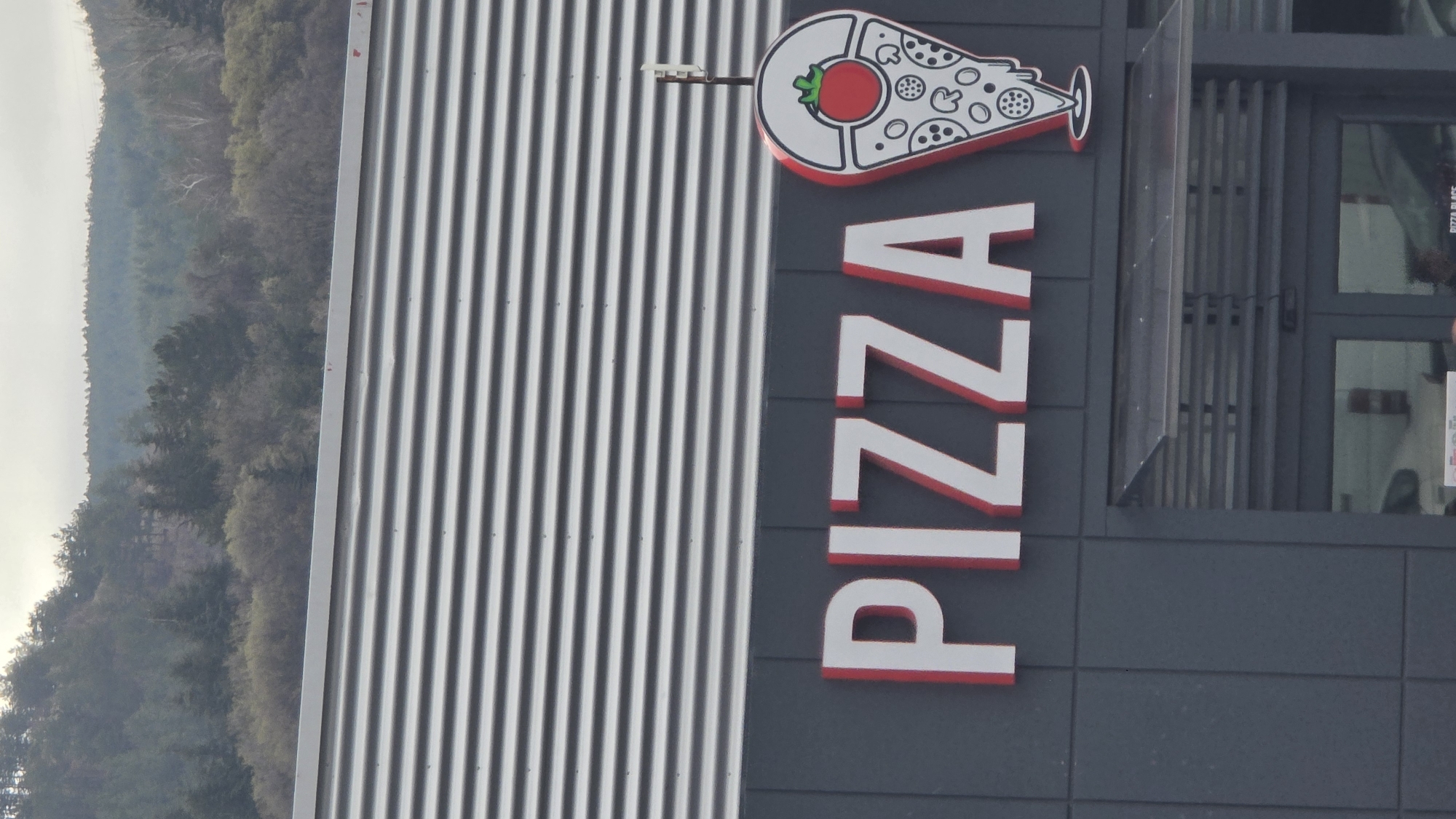 A slightly rotated medium shot shows a pizza restaurant. The sign reads "PIZZA", topped with a stylized pizza slice drawing. The restaurant's facade is a combination of corrugated metal and a dark gray tiled wall. A small security light is mounted above the sign. In the background to the left, there's a view of a tree-covered hillside under a hazy sky. Based on the sign, it is likely a Pizza Place in Blagoevgrad, Bulgaria. The doors can be seen to the right of the sign.