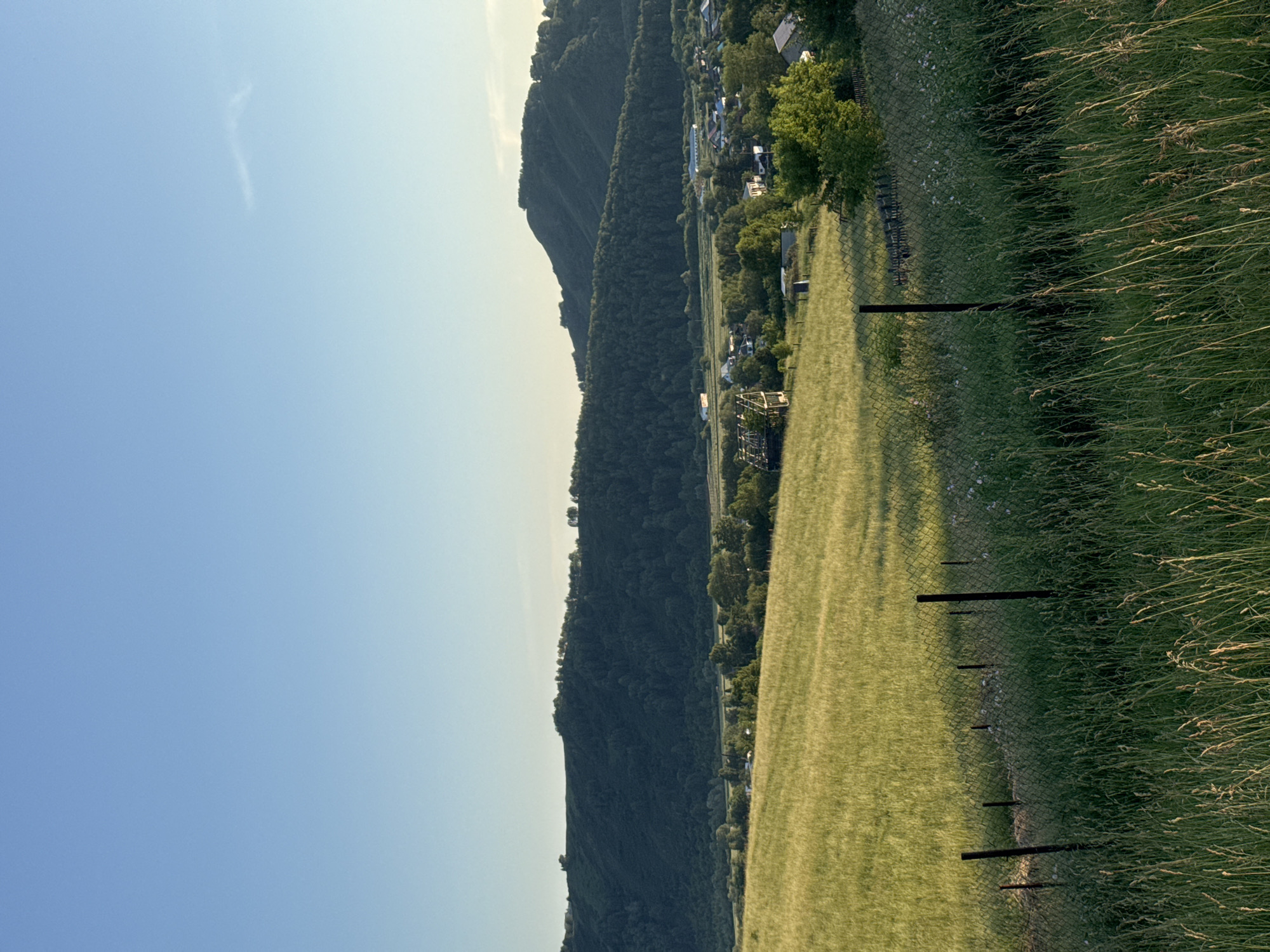 The image depicts a rural landscape in Soloneshnoye, Russia, likely during the late afternoon or early evening. The scene is dominated by a vast expanse of clear blue sky, gradually transitioning to a lighter hue near the horizon, suggesting a setting sun. A faint, wispy cloud is visible in the upper left portion of the sky.
The foreground features a gently sloping field of tall, golden-green grass, catching the warm light of the setting sun, which casts long shadows from several dark, vertical fence posts. A chain-link fence separates this grassy area from a slightly lower field of shorter, neatly mown grass, which appears to be a sports field or a park. In the distance, beyond the mown field, a small village is nestled at the base of a steep, densely forested mountain range. The houses appear to be spread out, surrounded by trees. The mountains are a deep, dark green, covered in a thick canopy of trees that obscure any details of the terrain. The overall impression is one of peace and tranquility, with the natural beauty of the landscape taking center stage. No people or specific activities are evident, but the presence of the village suggests a settled, rural community. The lighting and the long shadows indicate the golden hour of daylight, creating a serene and picturesque atmosphere.