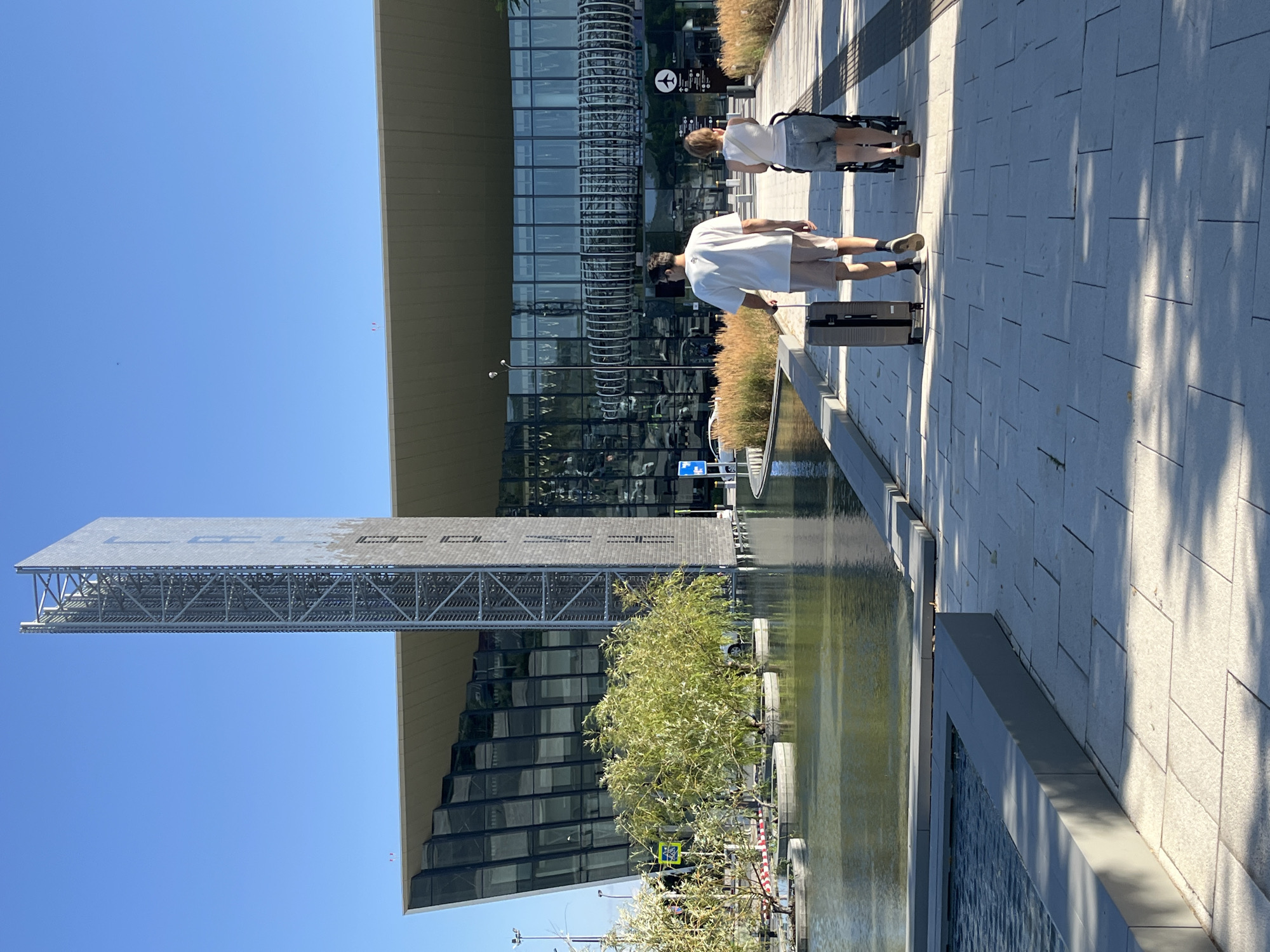 The image shows two people walking on a paved walkway in front of a modern building with a glass facade. The person in front is a man wearing a white t-shirt and beige shorts, pulling a rolling suitcase. The person behind him is a woman wearing a white tank top and denim shorts, pushing a stroller. To their left is a body of water with decorative trees planted in circular containers. Behind the building, a tall, metallic structure stands against a clear blue sky. The scene appears to be an outdoor area of an airport or a large public building. The location is identified as the city of Unknown, Russia.