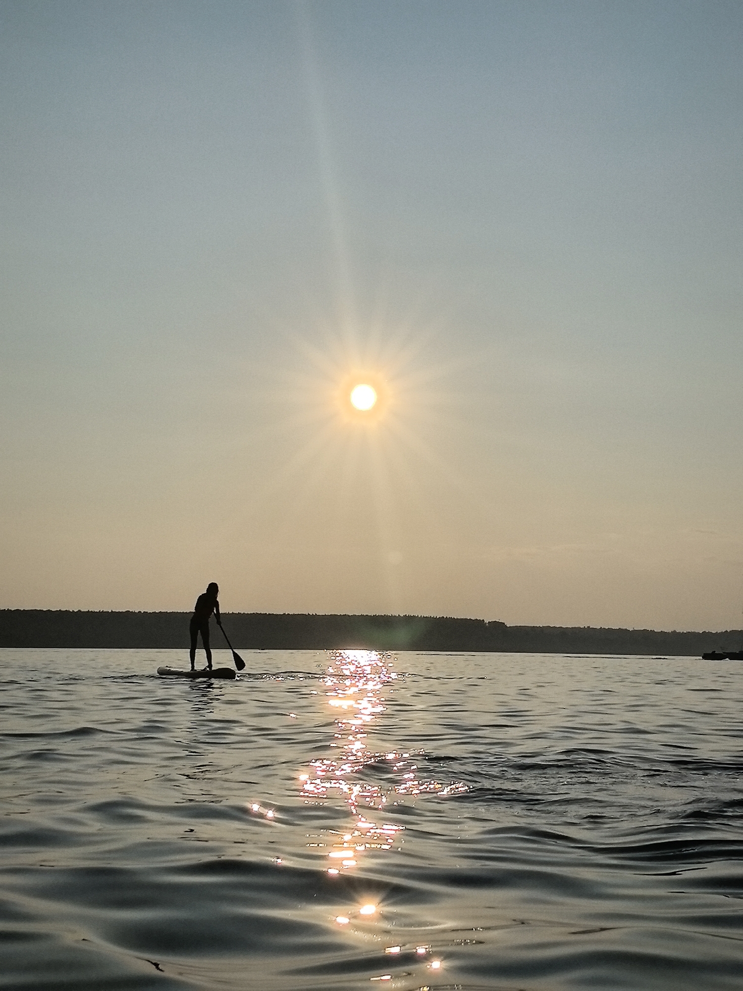 A lone person, silhouetted, is paddleboarding on a body of water. The sun is low in the sky, casting a bright glare and reflecting off the rippling surface of the water. A tree line is visible in the distance, forming the horizon. The sky is a pale blue, fading to a warmer yellow-orange near the sun. The scene appears to be during sunset or sunrise. It is not possible to determine the specific location in Perm, Russia from the image alone.