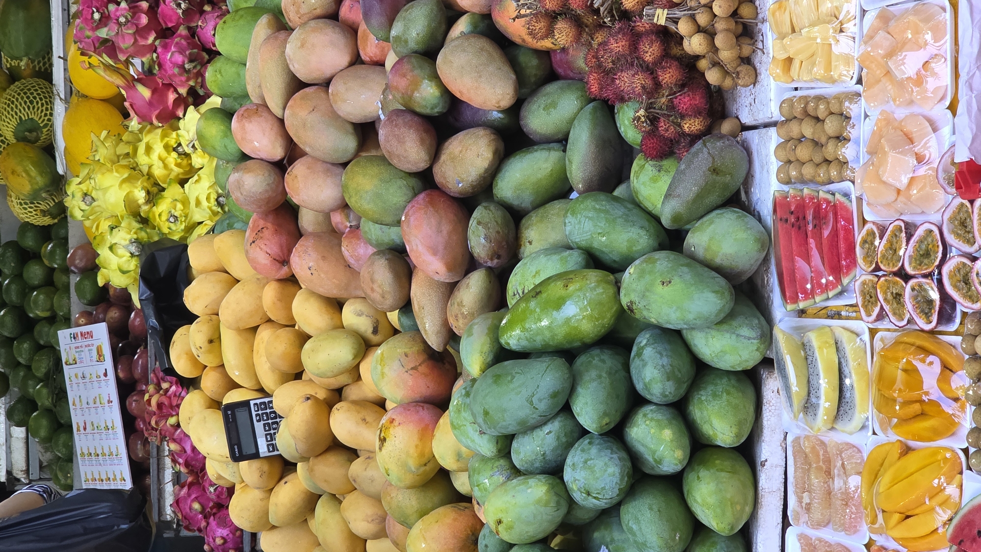 The image captures a vibrant fruit stand in Nha Trang, Vietnam. On display are various fruits like mangoes (in green, yellow and red varieties), dragon fruits (pink and yellow), rambutan, longan, limes, watermelon, passion fruit, durian, pomelo, and packaged sliced fruits. A calculator is seen atop a pile of mangoes, suggesting a transaction point. A menu with illustrations is positioned near the lime stack. The arrangement of fruits is dense, with a colorful display filling most of the frame.