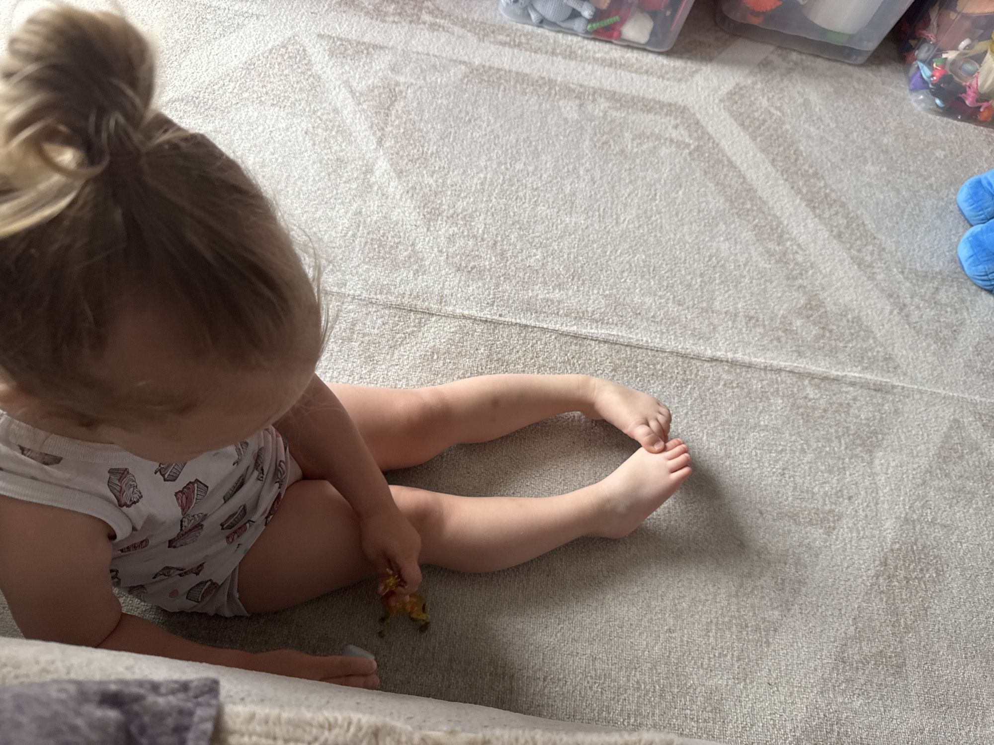 A young girl with blonde hair in a messy bun sits on a light-colored carpet with a geometric pattern. She is wearing white underwear with a repeating pattern of cupcakes. Her legs are extended in front of her, and her feet are bare. She is holding a small, colorful toy in her right hand, examining it. The carpet appears to be in a home setting, possibly a playroom, as evidenced by several clear storage bins filled with toys visible in the background. The lighting suggests it is daytime. The girl's posture and focus on the toy indicate she is engaged in play. There is no visible text in the image.