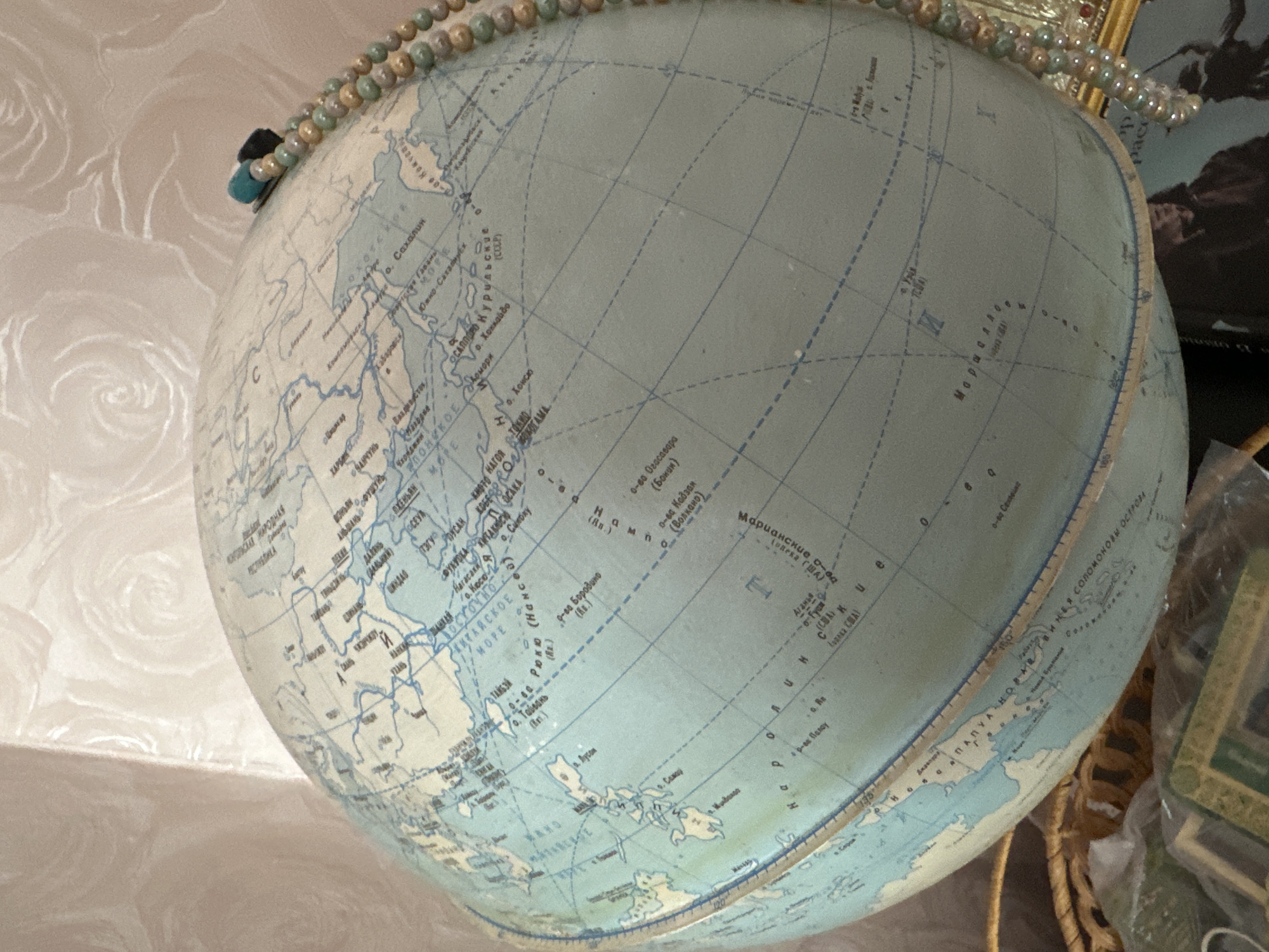 The image shows a close-up view of a vintage globe, presenting a portion of East Asia, including parts of Russia, Japan, the Korean Peninsula, and Southeast Asia. The globe appears to be an older model, with names written in Cyrillic and other languages. A beaded necklace is draped on the globe's top edge. The background reveals a patterned wall, a partial view of a framed picture, and what seems to be a woven basket holding other items. The exact location in Sayansk, Russia, cannot be determined.