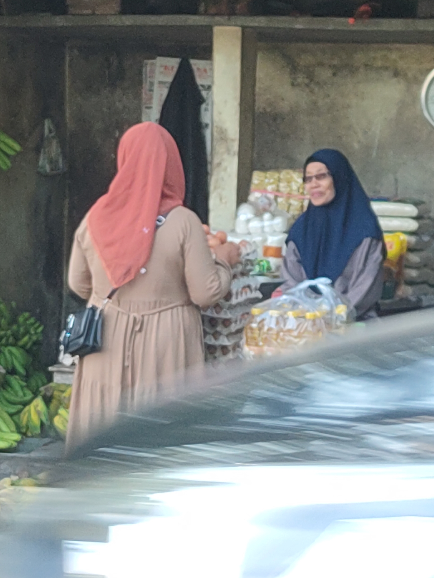 In Kediri City, Indonesia, two women are engaged in a transaction at an outdoor market stall. The woman on the left, wearing a reddish-orange hijab and a tan dress, has a black purse slung across her body. The woman on the right, wearing a dark blue hijab and a grey top, is interacting with the woman on the left. The stall displays various goods, including bananas on the left and what appear to be bags of rice and bottles of cooking oil on the right. The background shows a rough-textured wall and a shelf with items. The scene is captured in daylight, with a blurred foreground suggesting the photo was taken from a moving vehicle.