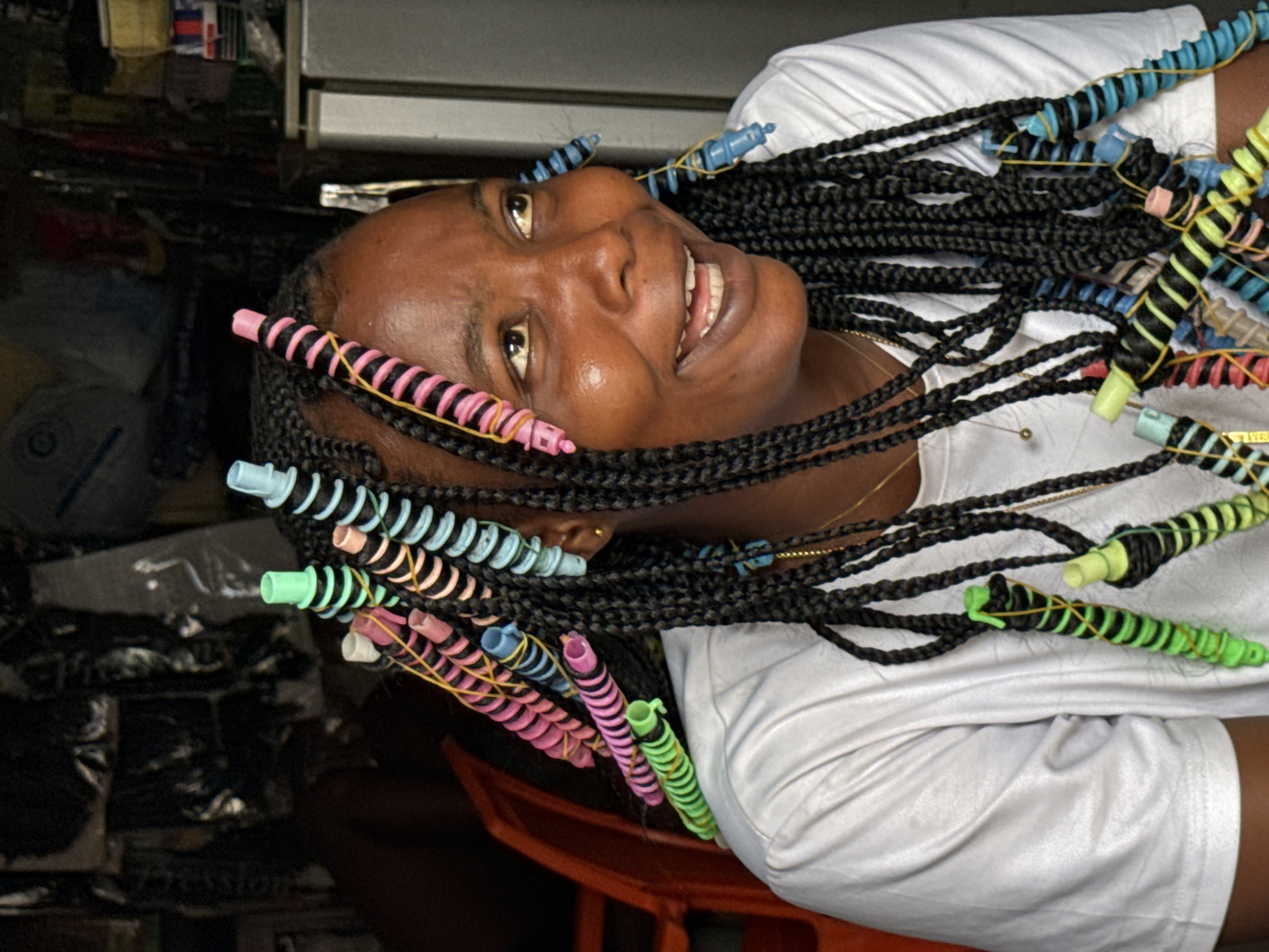 A portrait of a Black woman with braided hair adorned with colorful spiral curlers. She is wearing a white t-shirt and is smiling. The background appears to be a cluttered shop or storeroom in Ikoyi, Nigeria, with shelves filled with various items. The woman's face is illuminated, suggesting natural light.