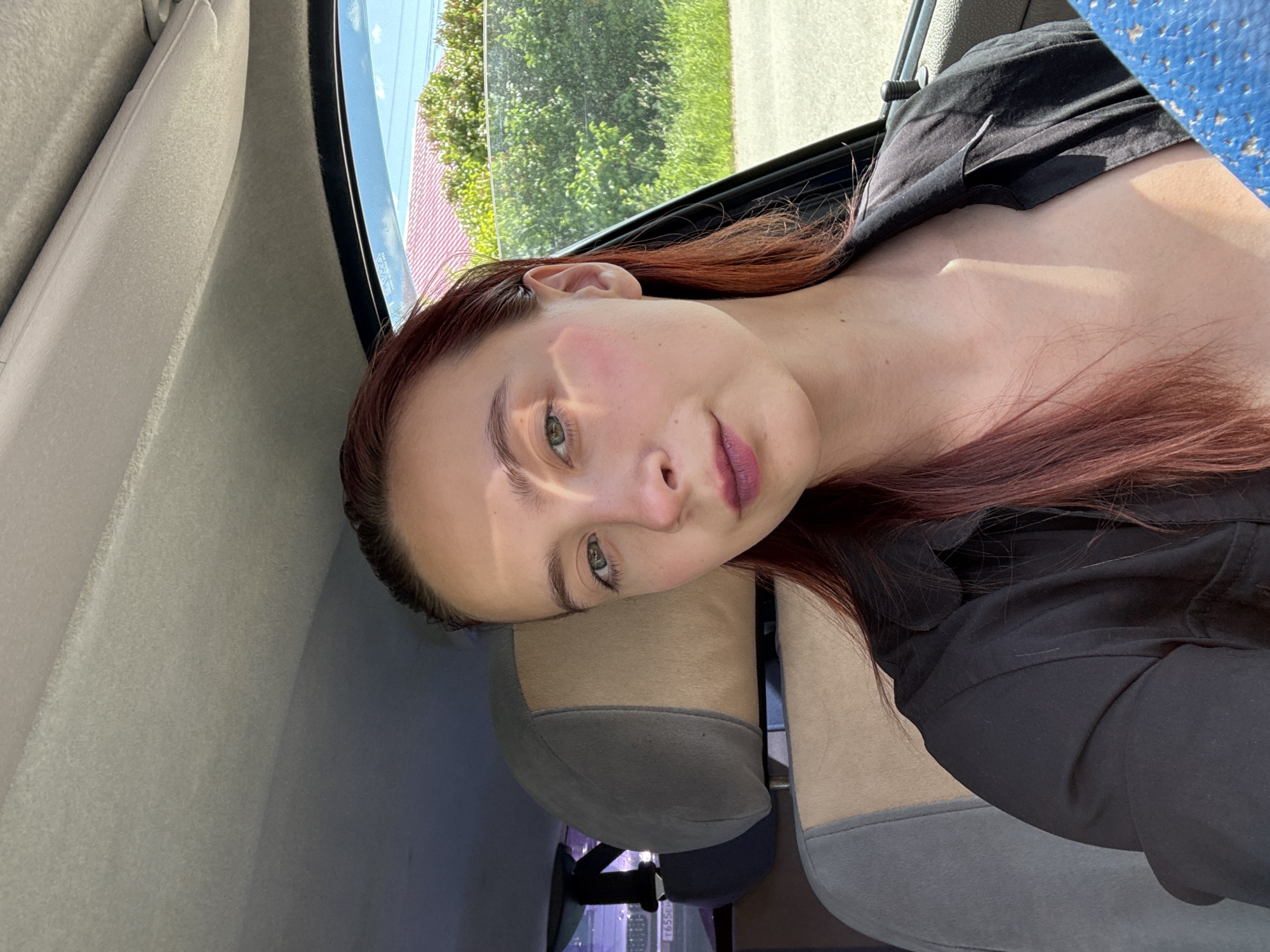 In the city of Perm, Russia, a young woman with red hair is taking a selfie inside a car. She is wearing a black shirt and her hair is falling around her shoulders. The interior of the car is visible, with a grey headrest and seat. Sunlight streams in from the open window, illuminating her face and neck. Outside the car, lush green trees and a red-tiled roof are visible under a bright blue sky. A license plate, partially obscured, is visible on the back of another vehicle in the background.
