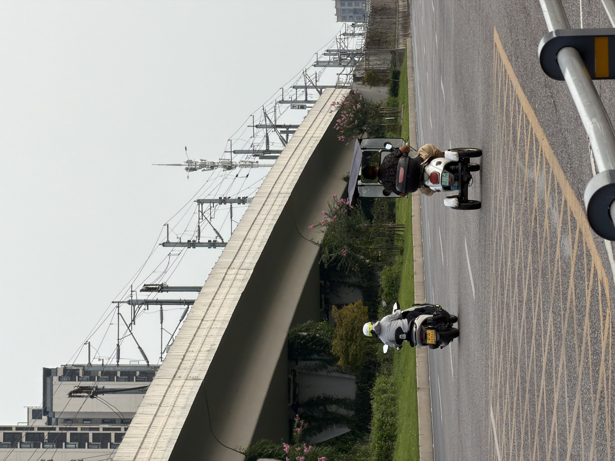 In Jiangjin, China, two individuals are seen riding motorized vehicles on a paved road under a large concrete overpass. The overpass is supported by pillars and appears to be part of a transportation infrastructure, possibly for trains, as evidenced by the overhead wires and poles. The sky is overcast.
On the left side of the road, a person wearing a white helmet and a light-colored jacket is riding a white scooter. The scooter has a yellow license plate that reads "88X66". Green grass and foliage, including some pink flowering bushes, are visible on the left side of the road, growing up the supports of the overpass.
On the right side of the road, another individual, wearing a dark patterned shirt and light-colored shorts, is riding a white three-wheeled scooter. This scooter has a canopy and a black storage box at the rear. The scooter's license plate is partially obscured.
The road surface is asphalt with yellow lane markings. In the foreground, yellow crisscrossing lines are painted on the road, and a gray metal barrier with yellow accents is visible on the right edge. In the background, modern buildings with many windows and communication towers can be seen.