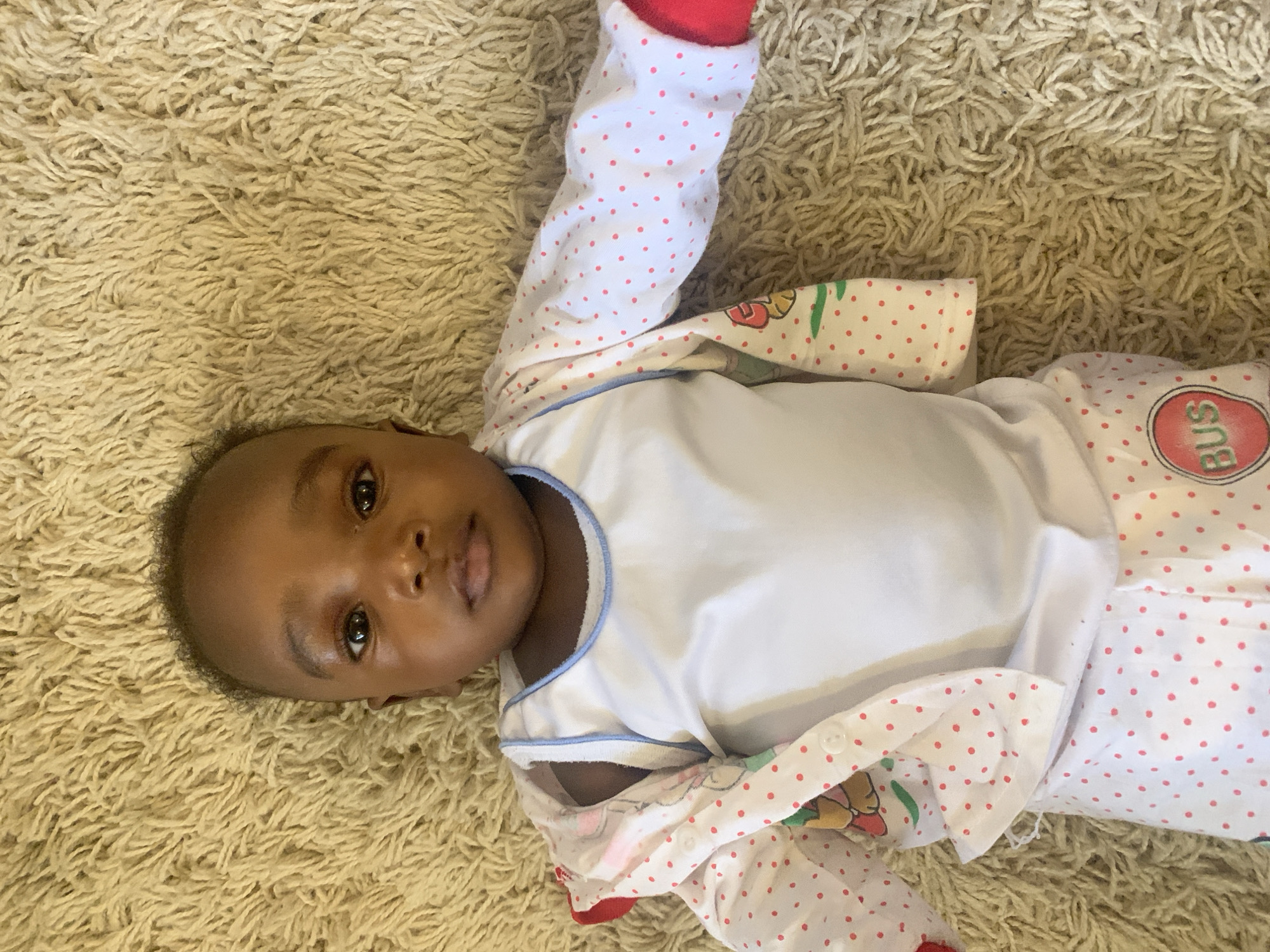 An infant is lying on their back on a light-colored, shaggy carpet in an indoor setting in Barakin Akawo, Nigeria. The baby, with dark skin and dark hair, looks directly at the camera with a calm, neutral expression. They are wearing a white undershirt with light blue trim around the neck and sleeves. Over the undershirt, the baby is dressed in a white garment, likely pajamas or a sleepsuit, adorned with small red polka dots. This outer garment also features colorful, cartoon-like illustrations and a prominent red circular patch on the right leg area, which clearly displays the word "BUS" in white letters. The lighting is soft, suggesting a relaxed daytime atmosphere indoors.