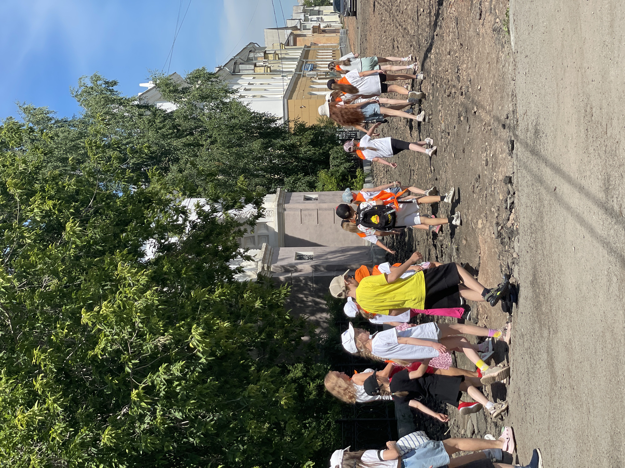 A group of children, some wearing orange neckerchiefs, are walking down a dirt road in an urban environment. They are accompanied by an adult woman with blonde hair. The road is made of dark, uneven stones. On the left side of the road, there is a black metal fence and lush green trees. Behind the trees, a white building with classical architectural details is visible. Further down the street, other buildings with light-colored facades line the road, and a car is parked on the right. The sky is clear and blue. The children appear to be on an outing or excursion.