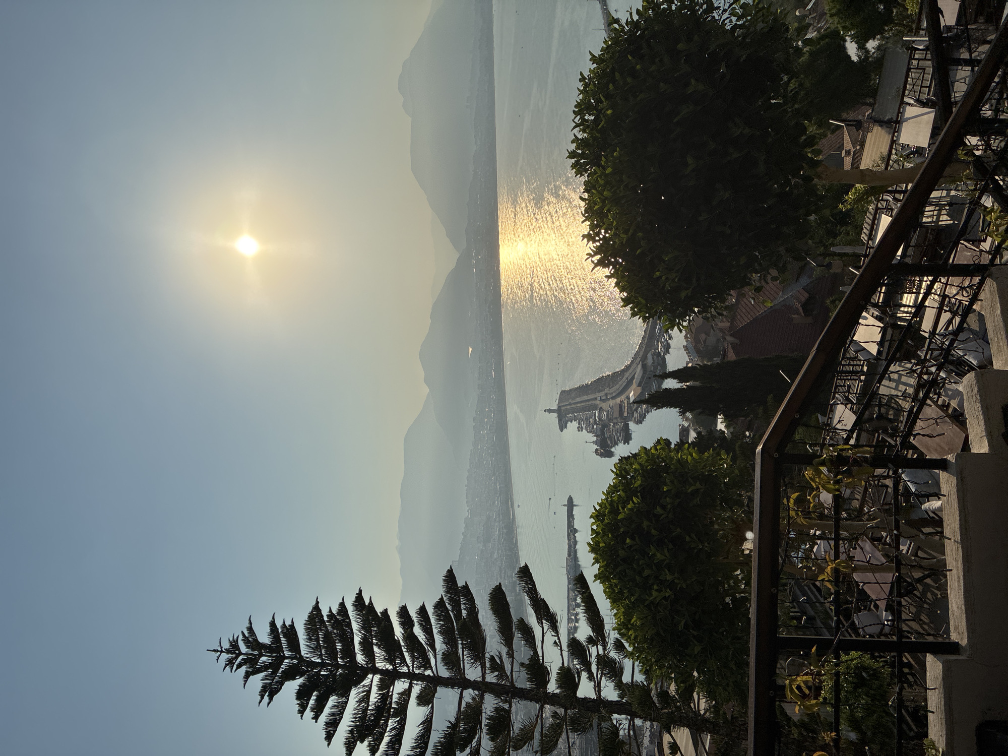 The image shows a scenic view of Alanya, Turkey, from an elevated vantage point, likely a terrace or balcony. The foreground features a railing and several tables and chairs, suggesting an outdoor seating area, possibly a restaurant or cafe. Two trees, one a tall, slender pine and another a fuller, rounded tree, are prominent on the left and right sides, respectively, framing the view.
In the middle ground, the city of Alanya spreads out along the coast, with buildings and structures visible. A prominent harbor juts out into the sea, with a lighthouse at its end. Numerous boats are docked in the harbor, and a few are visible on the water.
The background consists of the sea, which reflects the bright, hazy sunlight. The sun is high in the sky, creating a warm, golden glow on the water. Beyond the sea, a range of mountains form the horizon, their peaks softened by atmospheric haze. The sky is a pale blue, transitioning to a lighter hue near the horizon. The overall impression is one of a peaceful, sunny day in a coastal city.