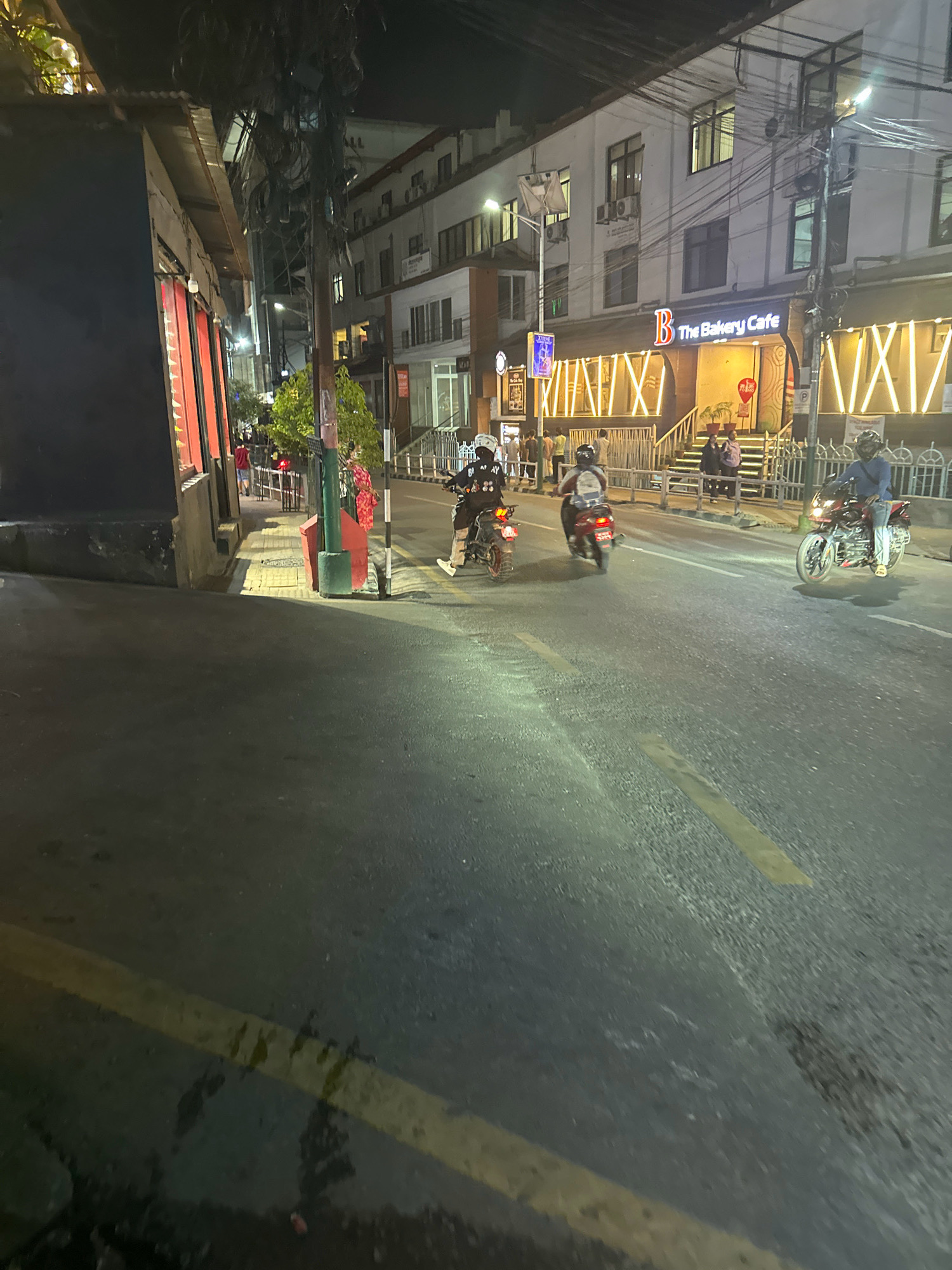 The image depicts a nighttime street scene in Kathmandu Metropolitan City, Nepal. A road with yellow lane markings occupies the foreground, with a sidewalk running parallel to it on the left. Several individuals are present, most notably three people on motorcycles. One motorcycle, a dark color with a red taillight, is in the foreground on the right, ridden by a person wearing a blue shirt and jeans, with a helmet. Another motorcycle, red, is further back on the right, carrying two individuals, both wearing helmets, one in a white shirt and the other in a darker top. A third motorcycle, darker in color, is further left and in the background, also with at least one person on it wearing a helmet.
On the sidewalk, there are several other people, some standing and some walking. To the right of the sidewalk is a building with "B The Bakery Cafe" prominently displayed on a lit sign. The building has multiple stories with windows and air conditioning units. Yellow decorative lights are installed along the facade of the cafe. On the left side of the image, a dark building with red-lit windows is partially visible. The overall environment suggests an urban setting at night, with artificial lighting illuminating the street and surrounding structures. The presence of motorcycles indicates vehicular traffic is active.