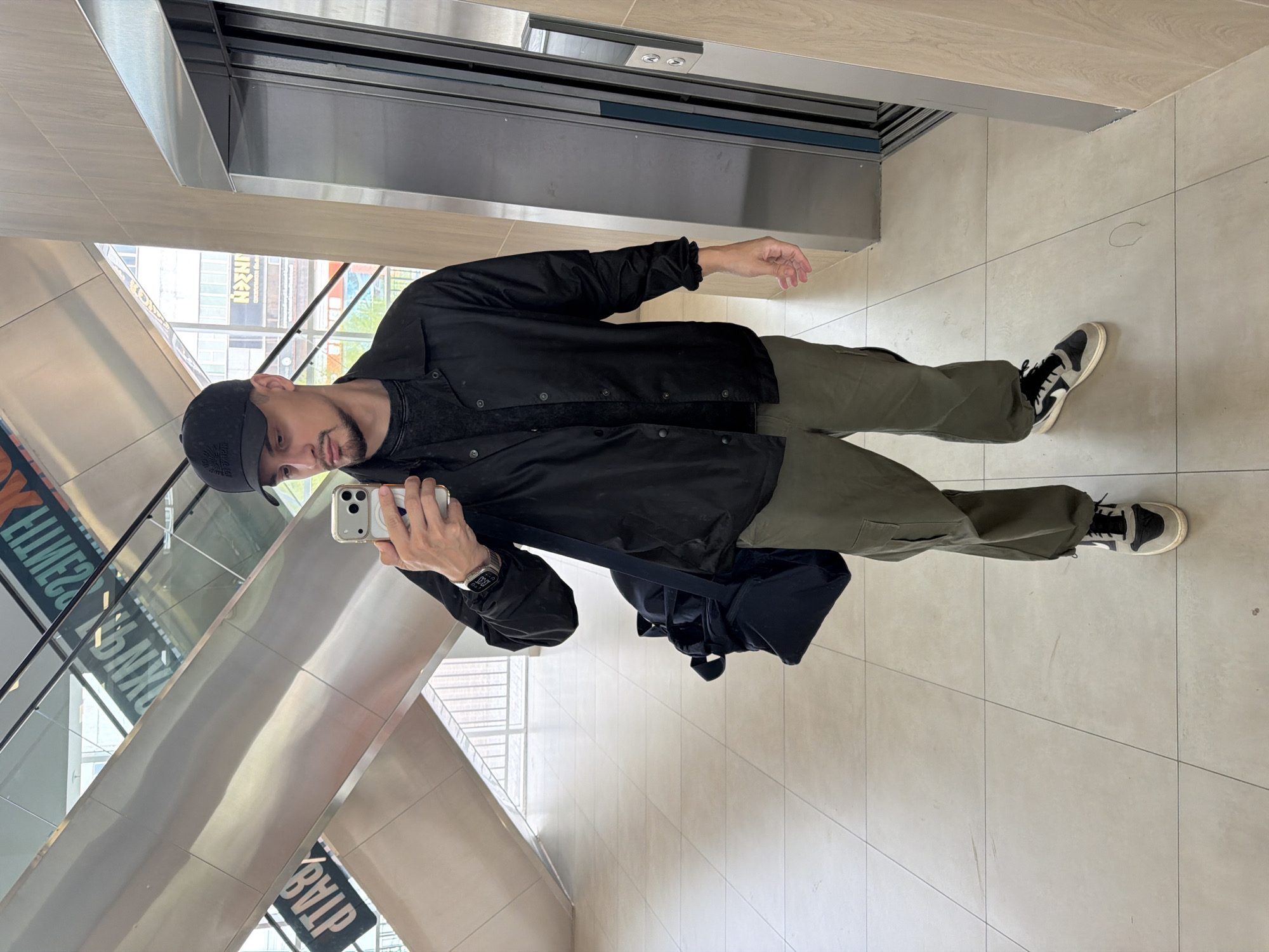 A man is taking a selfie in a building. He is wearing a black cap, a black jacket, green pants, and black and white sneakers. He is holding a silver iPhone in his right hand. A black backpack hangs over his left shoulder. The building appears to have an escalator and a metallic elevator. Reflected in the glass are signs with Cyrillic lettering, indicating the location could be in Krasnoyarsk, Russia.