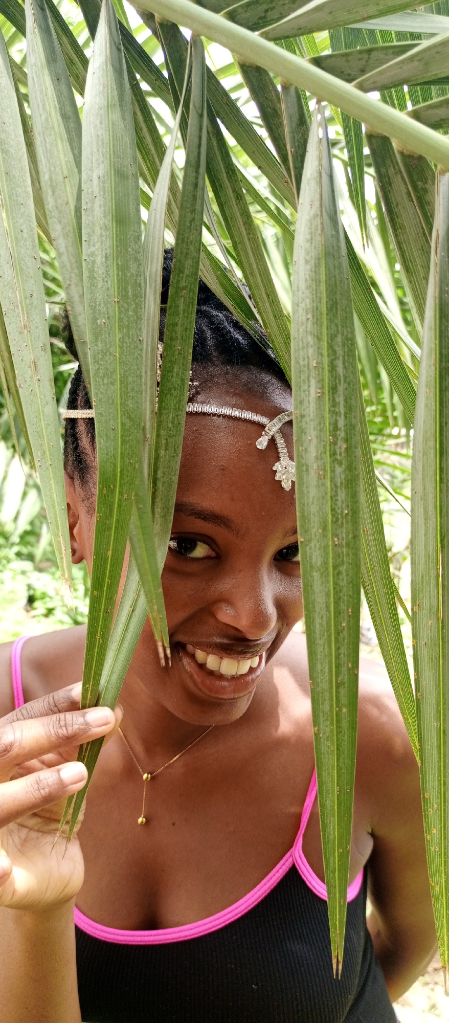 A young woman with dark skin, wearing a black top with pink straps and a bejeweled headpiece, smiles at the camera from behind several green palm leaves. She is holding one of the leaves in her left hand. She is also wearing a delicate gold necklace. The background is blurred green foliage. The location is Keffi, Nigeria.