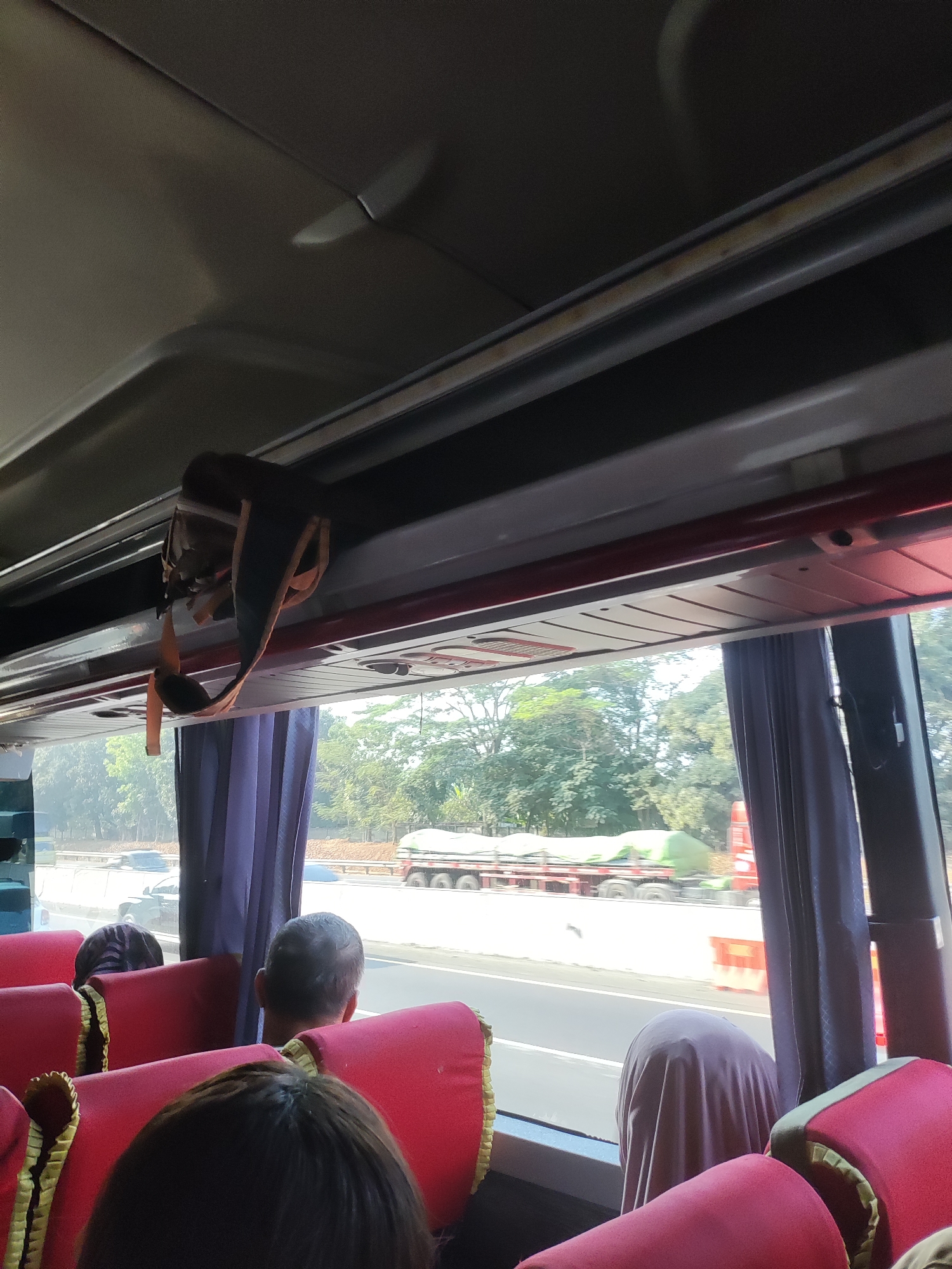 This image, taken from inside a bus, shows a view of the Karawang region of Indonesia. The interior of the bus features red seats, some of which are occupied by passengers. A strap hangs from the ceiling of the bus. Outside the bus window, a highway is visible with several vehicles, including cars and a truck carrying a green tarped load. Trees line the side of the highway, indicating a semi-rural or suburban setting. The sky is bright, suggesting daylight.