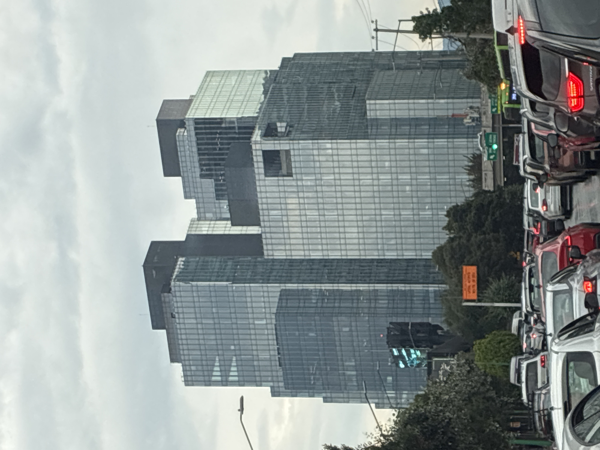 The image shows a dense urban scene, likely in Mexico City, Mexico, based on the architectural style of the buildings. The dominant feature is a large, modern glass skyscraper with several distinct levels or sections, giving it a layered appearance. A significant traffic jam is visible on a street adjacent to the skyscraper, with numerous cars and buses closely packed together, suggesting a high volume of vehicular traffic. The sky is overcast and cloudy, which might suggest a rainy or humid day. The photograph is taken from a slightly oblique angle, giving the impression of being shot from within another vehicle stuck in the same traffic jam. Streetlights, traffic signals, and some greenery are present, adding further details to the urban environment.