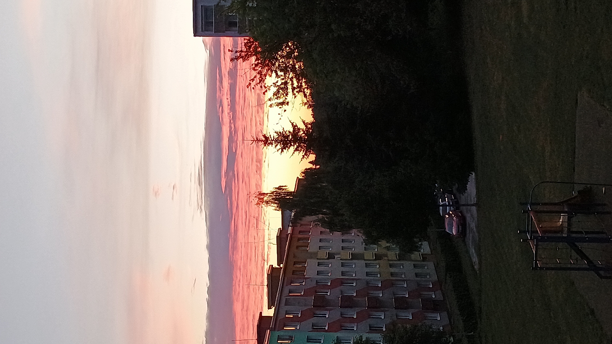 A vertical, medium shot captures a view of a residential area at dusk in Sokolov, Czechia. In the foreground, a dark, grassy playground area is visible, featuring a small, blue and red climbing structure with a slide. In the mid-ground, a line of dark green trees forms a dense border. Beyond the trees, a row of apartment buildings runs horizontally across the frame. The buildings are multi-story and have a colorful, patterned facade with many windows. Several parked cars are visible in front of the buildings, including a red car. The sky above is a gradient of soft blues and pinks, with horizontal bands of clouds reflecting the sunset. The overall scene depicts a tranquil evening in a city neighborhood.