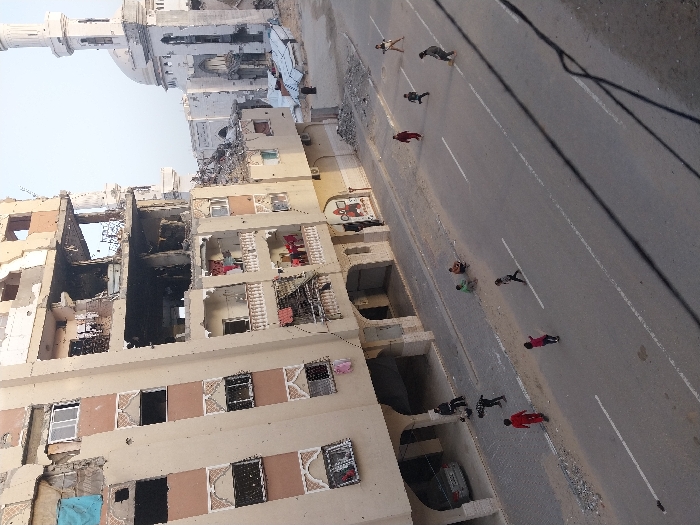 An aerial view captures a street scene in Khan Yunis, Palestinian Territory, presenting a striking contrast between urban decay and daily life. On the left, a multi-story building complex stands severely damaged, with large sections of its walls and floors missing, exposing interiors and showing signs of fire damage with blackened areas and visible debris. Windows are broken or absent. In the upper background, a mosque with a tall minaret and a prominent dome is visible.
The wide asphalt road, marked with white lane lines, stretches across the right side of the image. Several dark utility cables hang overhead, casting distinct shadows on the street. Scattered across the road are approximately ten individuals, mostly appearing to be children or young adults, dressed in casual clothing of various colors such as red, green, and black. Some are standing or walking, while others appear to be sitting or lying on the pavement. A car is partially visible, parked beneath an archway of the damaged building.
The scene is set during the daytime, indicated by the bright lighting and clear shadows. The extensive destruction of the buildings suggests the aftermath of conflict or a major incident. No discernible text is visible.