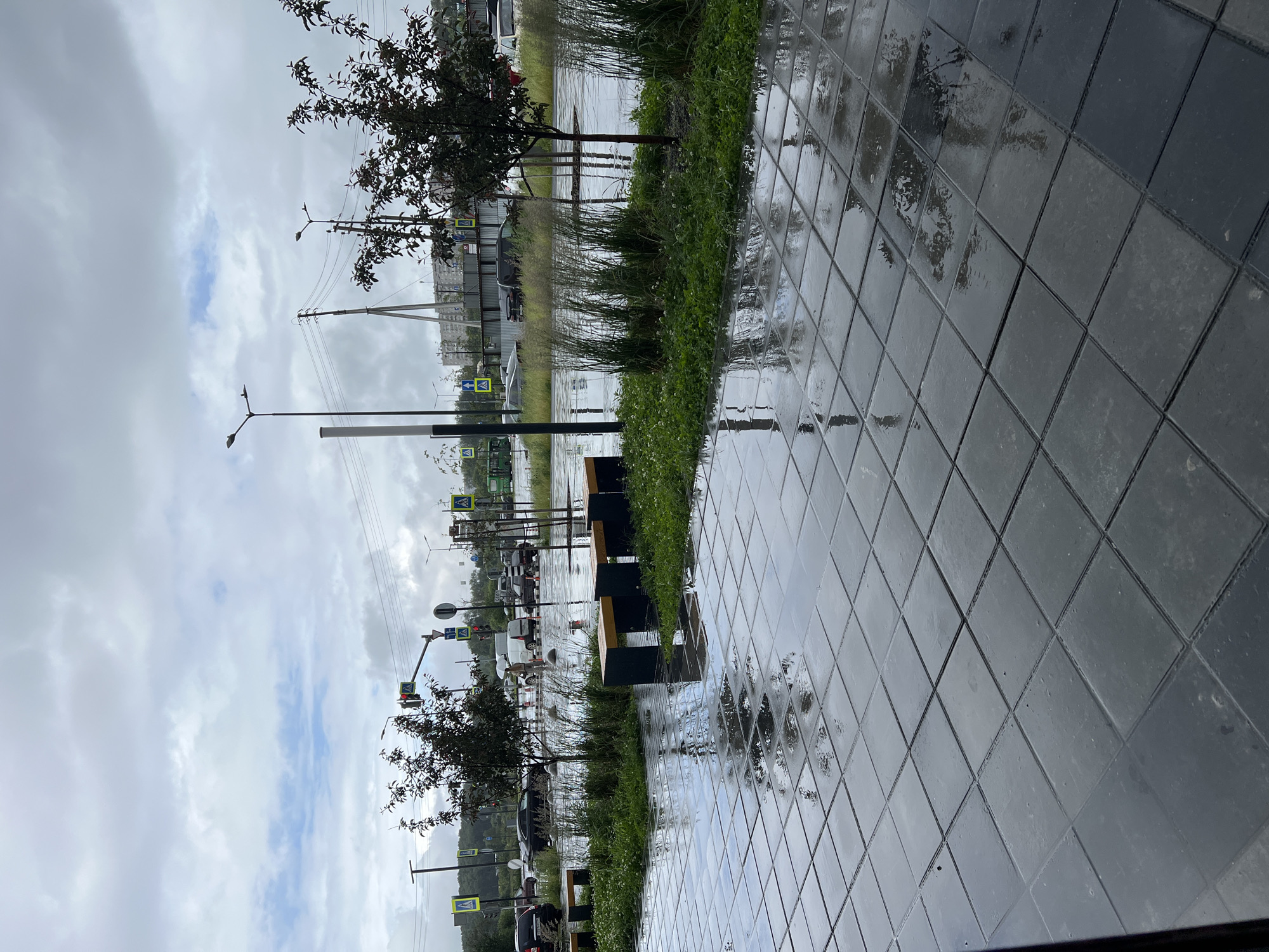 The image depicts a wet, paved urban environment, likely Yekaterinburg, Russia, during overcast weather. The foreground is dominated by dark gray, square paving stones that are wet and reflective, showing a distorted image of the sky and surrounding elements. A line of low green plants and tall ornamental grasses runs horizontally across the mid-ground, bordered by a wet, reflective surface that appears to be a shallow pool or very wet pavement. Several dark benches with wooden seats are situated along this grassy area.
Beyond the vegetation, a road is visible with several vehicles, including cars and a white van. A traffic light shows a red signal, indicating a stop. Pedestrian crossing signs with yellow triangular backgrounds and a black pedestrian silhouette are present near the traffic signal. Streetlights are also visible. In the background, there are buildings and construction elements, including what appears to be a bridge or overpass structure. The sky is heavily clouded, contributing to the muted light and overall damp atmosphere. The scene appears to be an outdoor public space, possibly a park or a pedestrian area adjacent to a road.