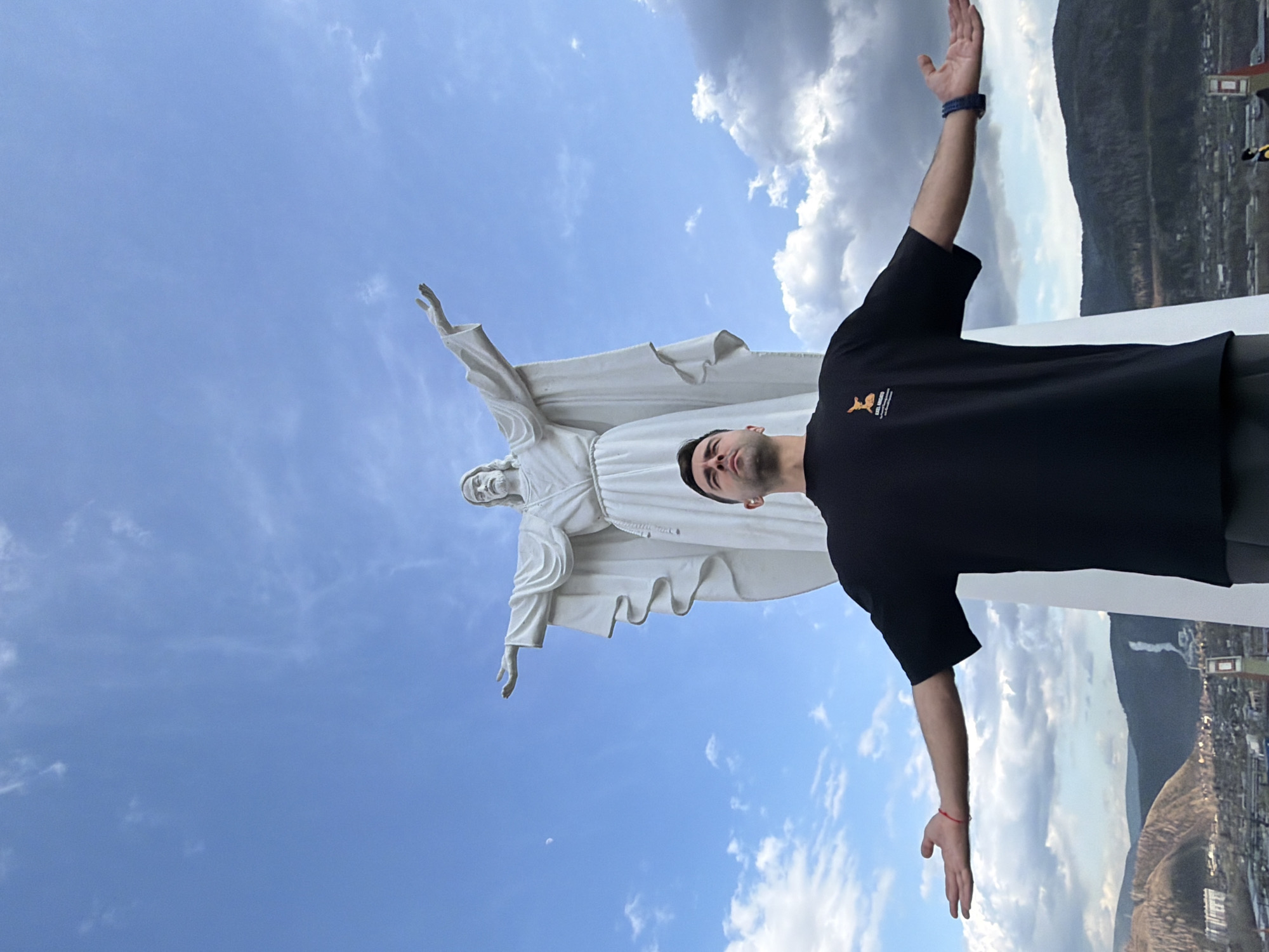 A man in a black t-shirt stands with his arms outstretched, mirroring the pose of the Christ the Redeemer statue in Cochabamba, Bolivia. The sky is partly cloudy. The city and mountains are visible in the background. The statue is white. The man is wearing a bracelet.
