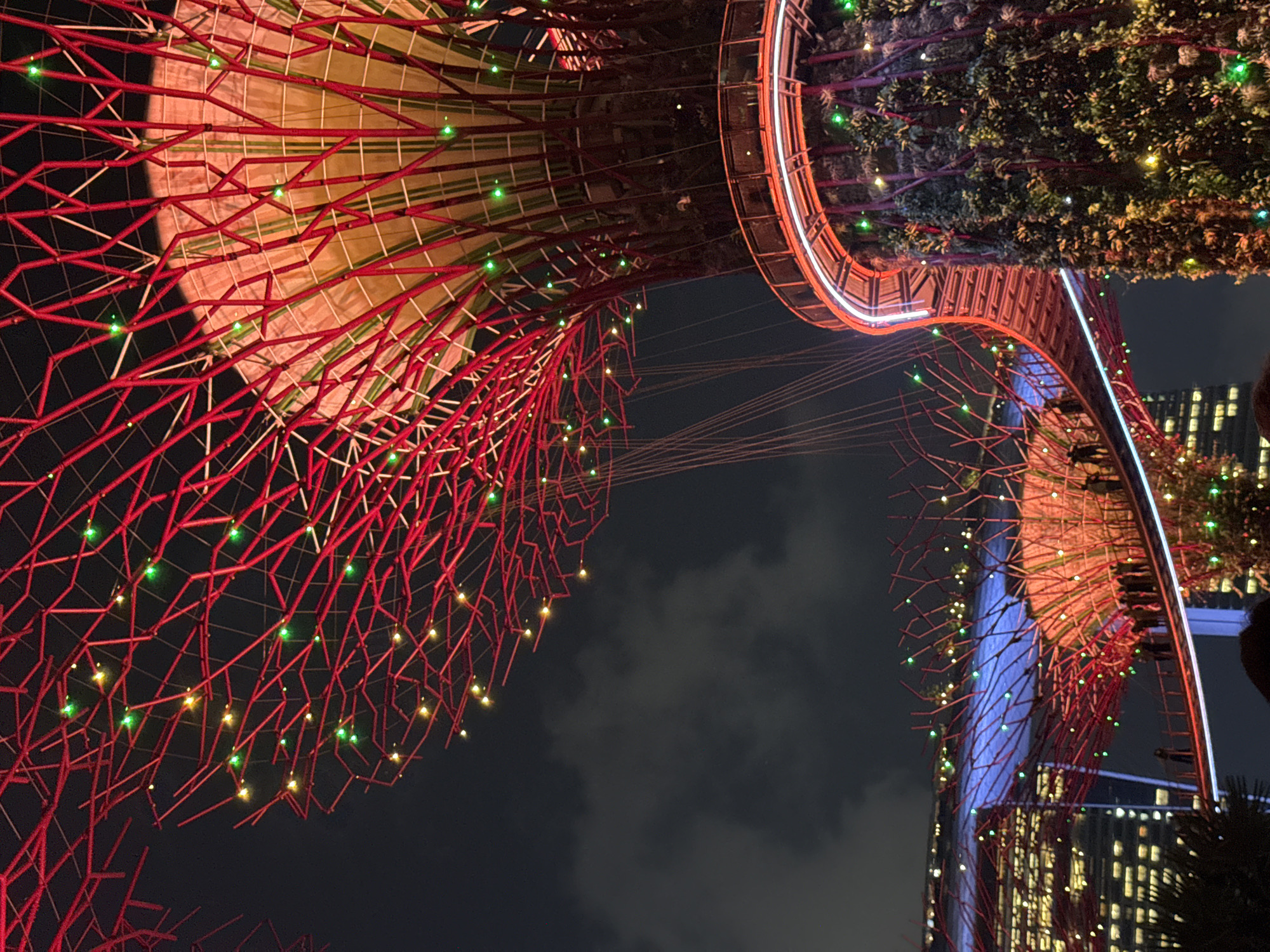 This is a nighttime photograph of the Supertree Grove at Gardens by the Bay in Singapore. The foreground features a massive, tree-like structure with a vibrant red lattice canopy adorned with green and yellow lights. A curved, illuminated walkway is visible on the right, wrapping around a portion of the structure which is covered in lush greenery. In the mid-ground, another similar structure is seen with a red lattice canopy and lights, connected by a skywalk. Silhouettes of people can be observed walking on this skywalk. To the left, a modern building with illuminated windows is visible, hinting at the urban setting. The sky is dark and cloudy, with some diffused lighting.