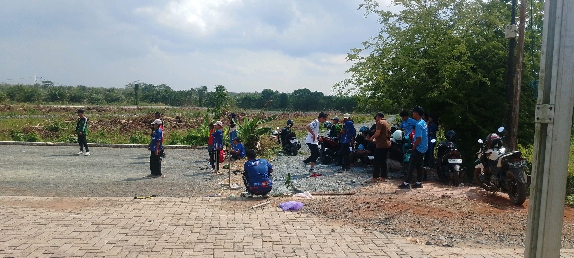 The image depicts an outdoor scene with a group of people gathered near motorcycles. The environment appears to be a semi-developed area, possibly on the outskirts of Batulicin, Indonesia, with cleared land, vegetation, and a dirt/gravel road. Several young men and women are present, some standing, some sitting, and some interacting with each other. Two motorcycles are parked side-by-side, and another is visible further to the left. The people are dressed in casual clothing, with some wearing blue shirts. The sky is partly cloudy. In the foreground, there is a paved brick area. The overall impression is a casual gathering or break in an outdoor setting.