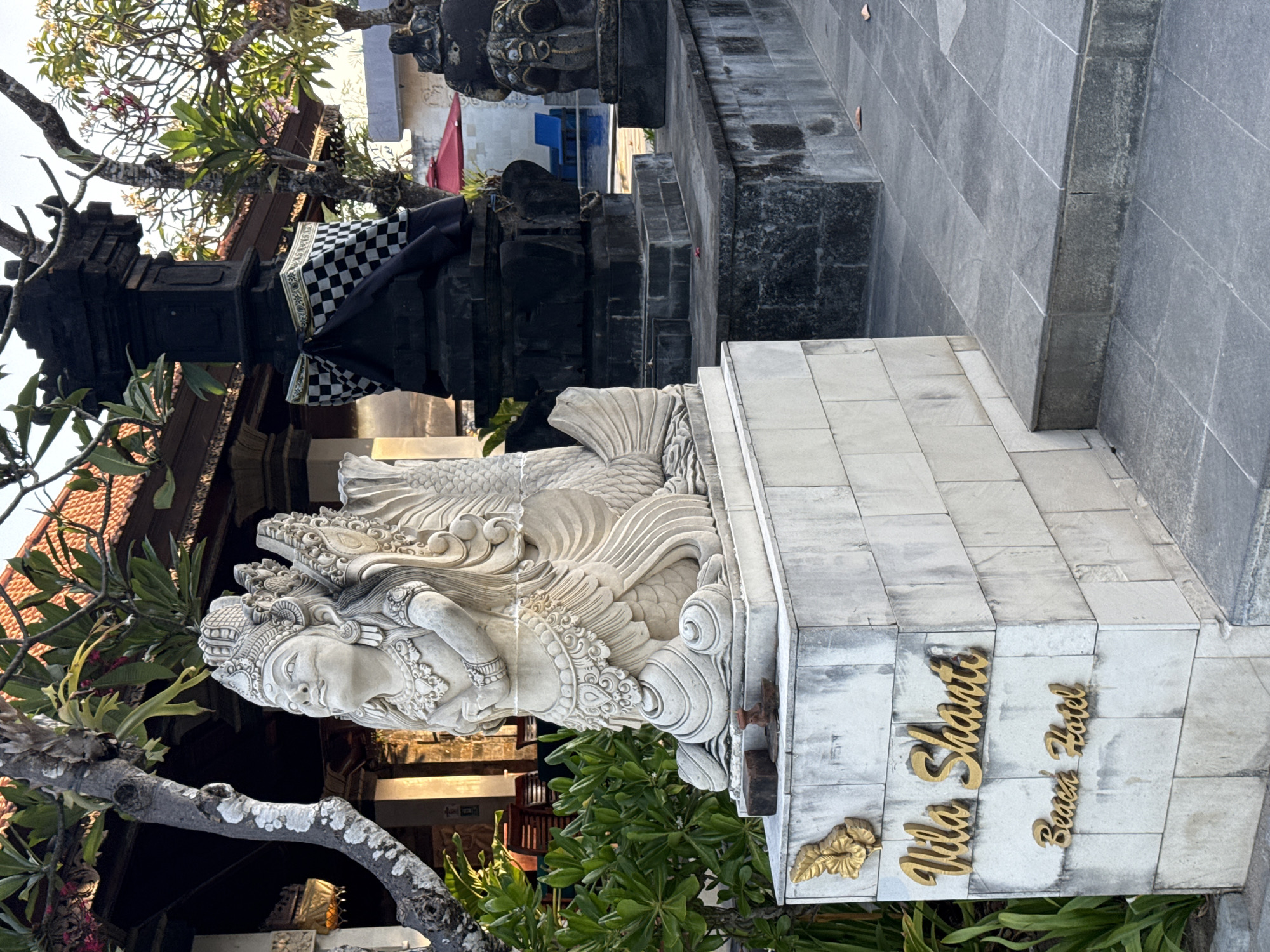 The image shows a stone statue of a mythological creature in front of a building. The statue is white and depicts a human torso with wings and a fish tail, kneeling on a stone base. The base has golden lettering that reads "Vila Shanti Beach Hotel". To the right of the statue is a dark stone monument adorned with a black and white checkered cloth. In the background, a building with a tiled roof and trees with green foliage can be seen. The ground is paved with grey tiles and stones. The scene appears to be an entrance to a hotel or resort. This location is Vila Shanti Beach Hotel, Denpasar, Indonesia.