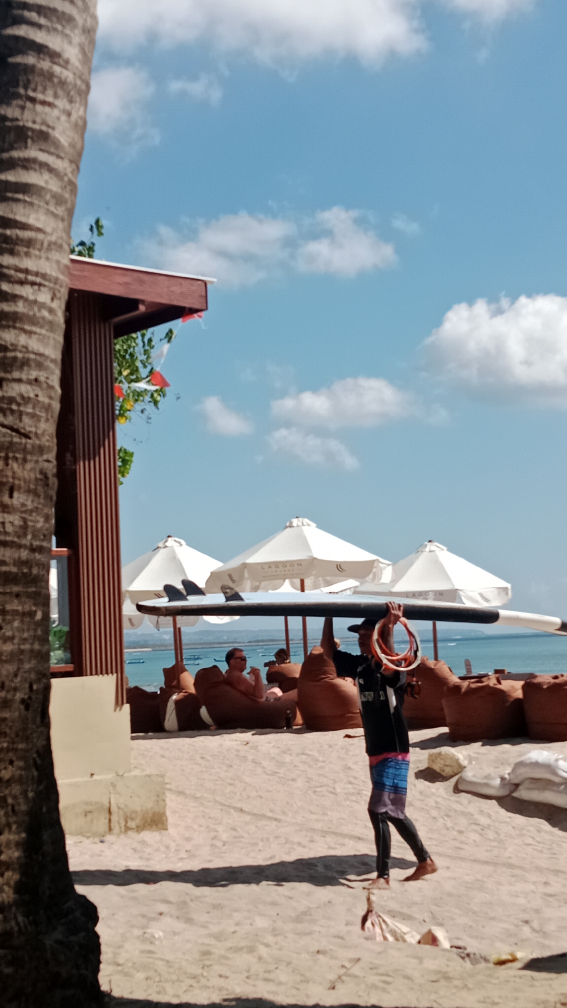 A man walks across a sandy beach in Kuta, Indonesia, carrying a surfboard balanced on his head. He wears a black long-sleeved shirt, colorful patterned shorts, and black leggings. In his right hand, he holds several orange and red coiled ropes. The beach is dotted with large, brown beanbag chairs, some occupied by beachgoers relaxing under white umbrellas. In the background, the ocean is visible under a bright blue sky with scattered white clouds. A palm tree trunk is prominent in the left foreground, partially obscuring a section of a brown wooden structure.
