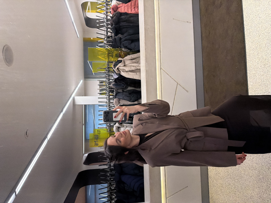 A woman in a belted jacket poses for a mirror selfie. She holds her phone in her right hand, while wearing a dark dress. The mirror reflects a bright space with a coat rack holding many coats. The floor transitions from brown carpet to a lighter, speckled material. The mirror itself has geometric designs.