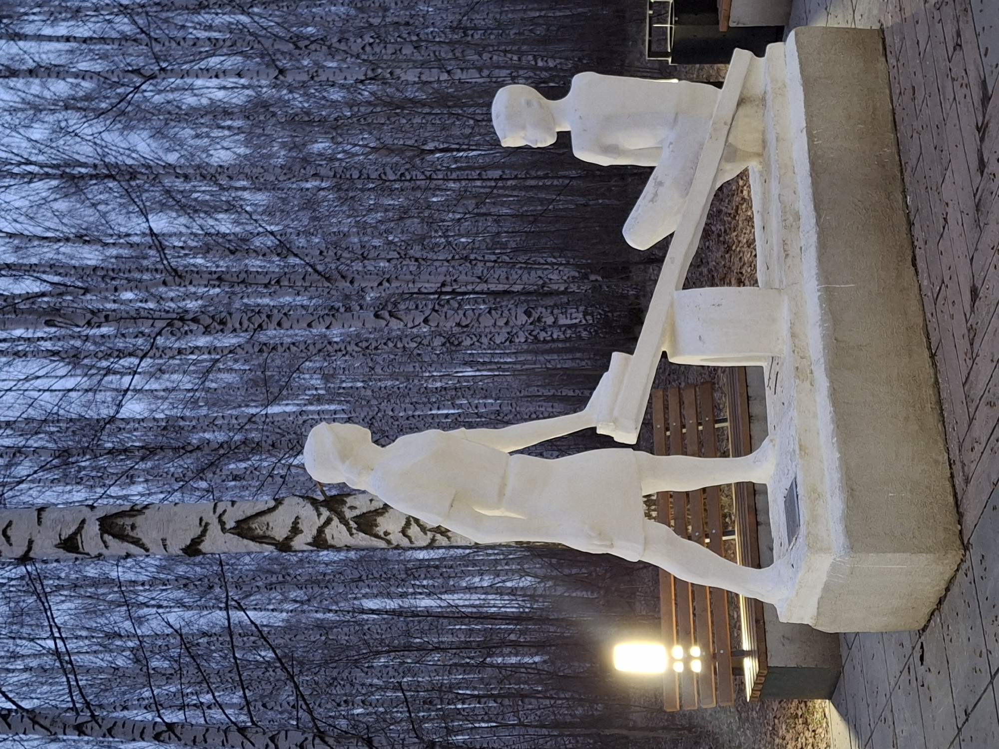 The image depicts a white, minimalist sculpture of two figures standing before a desk, likely a public artwork. The sculpture is placed on a concrete plinth, beside a park bench and a brick paved area. The background consists of a dense birch tree forest with barren branches against a blue-grey sky, indicative of either winter or late autumn. The sculpture is identified as being located in the city of Chaikovsky, Russia. A light source is visible near the bench illuminating the lower part of the scene.