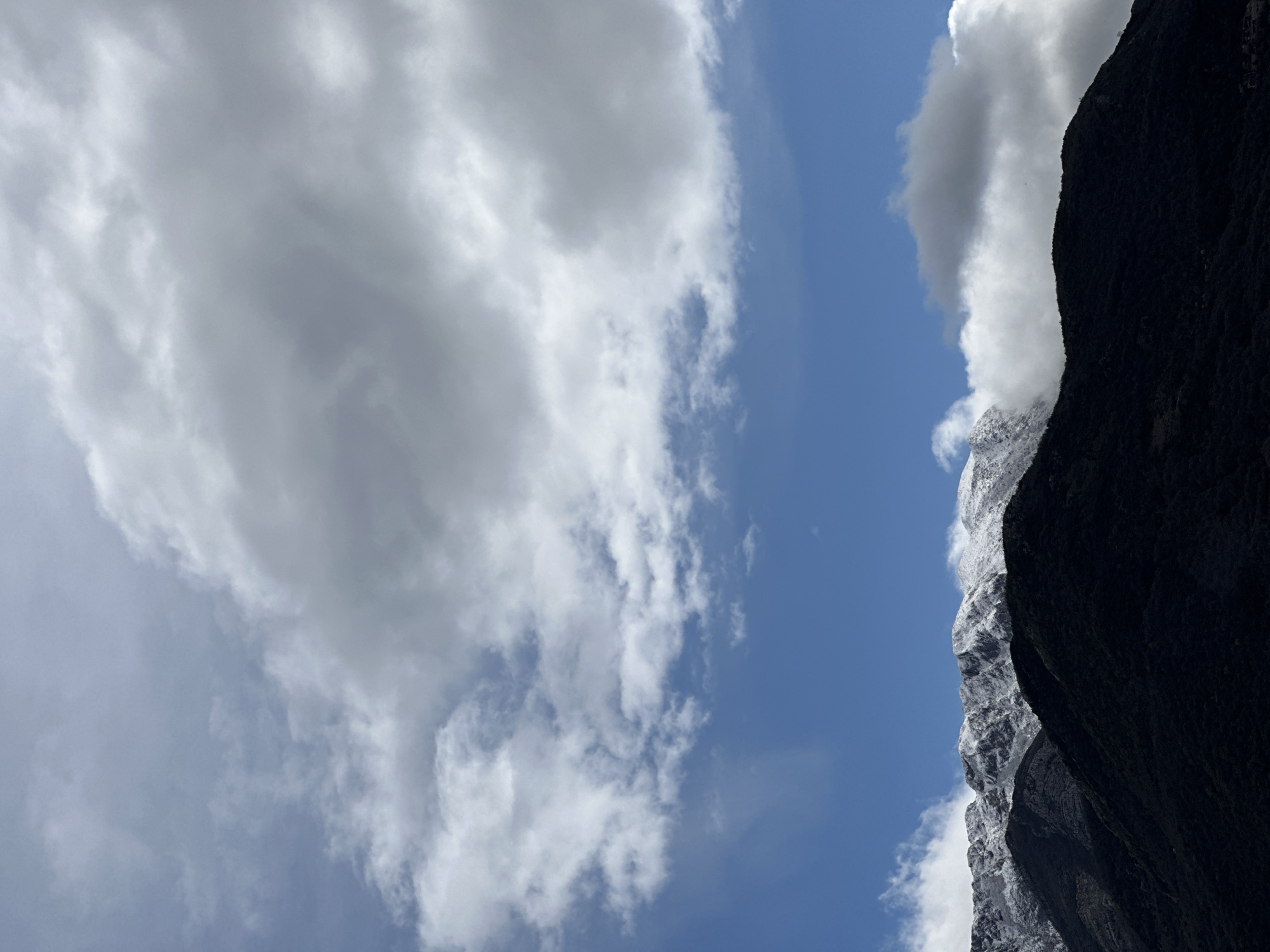 The image displays a mountainous landscape under a partially cloudy sky, observed from the vicinity of Šušanj, Montenegro. The lower portion of the frame features dark, tree-covered mountain slopes, exhibiting varied textures from dense foliage to exposed rock faces. Above these, the upper mountain peaks are visible, uniformly covered with a layer of white snow. Clouds are present throughout the sky: dense, grey stratocumulus clouds occupy the upper left and upper right quadrants, transitioning to lighter, whiter cumulus clouds along the mid-horizon, partially obscuring the snow-capped peaks. A clear, vibrant blue sky is visible in the middle section of the frame, separating the upper cloud layer from the mountainous terrain. No people, animals, or man-made structures are discernible within the frame. The lighting suggests an overcast yet bright condition, highlighting the snow against the darker slopes.