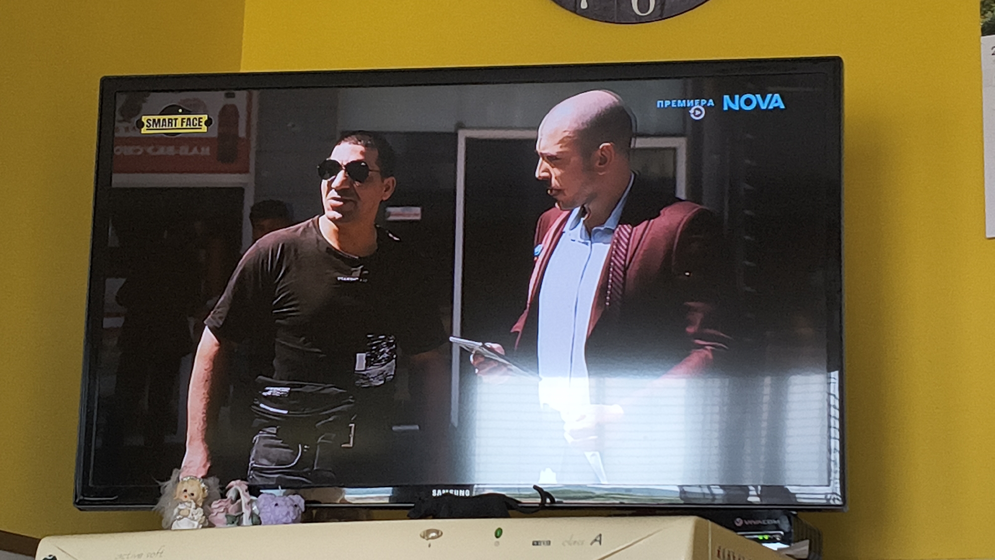 The scene depicts an indoor environment in the city of Shumen, Bulgaria, featuring a flat-screen television displaying a broadcast. The television, identified by a "SAMSUNG" logo on its bottom bezel, rests on a light-colored piece of furniture.
The TV screen shows two adult males. The man on the left has a dark complexion, wears dark sunglasses, a black short-sleeved t-shirt, and a black utility belt or fanny pack around his waist. He is facing slightly to his right. The man on the right is bald, has a lighter complexion, and is dressed in a light blue collared shirt underneath a maroon jacket with distinct vertical stripes on the shoulders. He holds a white, rectangular object, possibly a tablet or a book, in his hands and is looking towards the man on the left.
On-screen graphics include a yellow rectangular logo with "SMART FACE" written in black text in the top-left corner. In the top-right corner, the Bulgarian word "ПРЕМИЕРА" (PREMIERE) is displayed next to the "NOVA" television channel logo. The lower half of the screen exhibits prominent vertical light reflections, indicative of blinds or a window.
In the foreground, directly below the television, are several small decorative objects: a small doll dressed as a bride, a miniature woven basket with floral embellishments, and a small purple figurine. A black cable or strap is visible near the bottom center of the TV bezel. To the right of these objects, beneath the TV, is a black electronic device labeled "WINCOM".
The wall behind and to the right of the television is painted a solid yellow. A partial circular object, possibly a clock or decoration, is visible on the yellow wall above the TV, and a white, rectangular piece of paper, possibly a calendar, is partially visible on the wall to the far right.