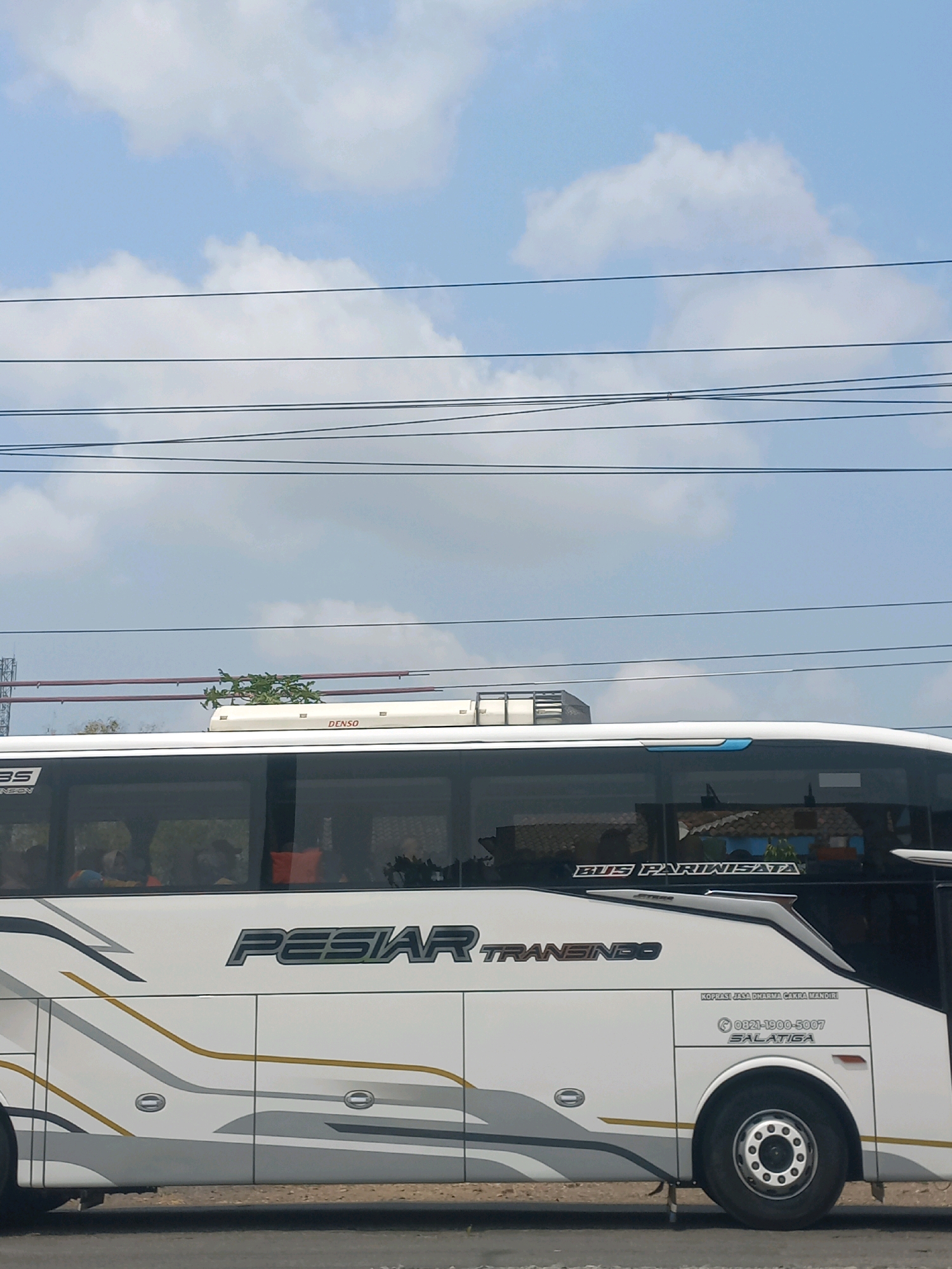 The image shows the side of a white bus with "PESIAR TRANSINDO" written on it in large letters. The bus is traveling on a road, with power lines and a cloudy blue sky visible above. The "DENSO" logo is visible on the roof of the bus. The text "BUS PARIWISATA" is visible on the side of the bus, indicating it is a tourist bus. Below that, there is contact information for "KOPIASI JASA DHARMA CAKRA MANDIRI" in Salatiga, Indonesia. The scene is likely in Baturetno, Indonesia, based on the provided location. People are visible through the tinted windows of the bus, appearing to be passengers.