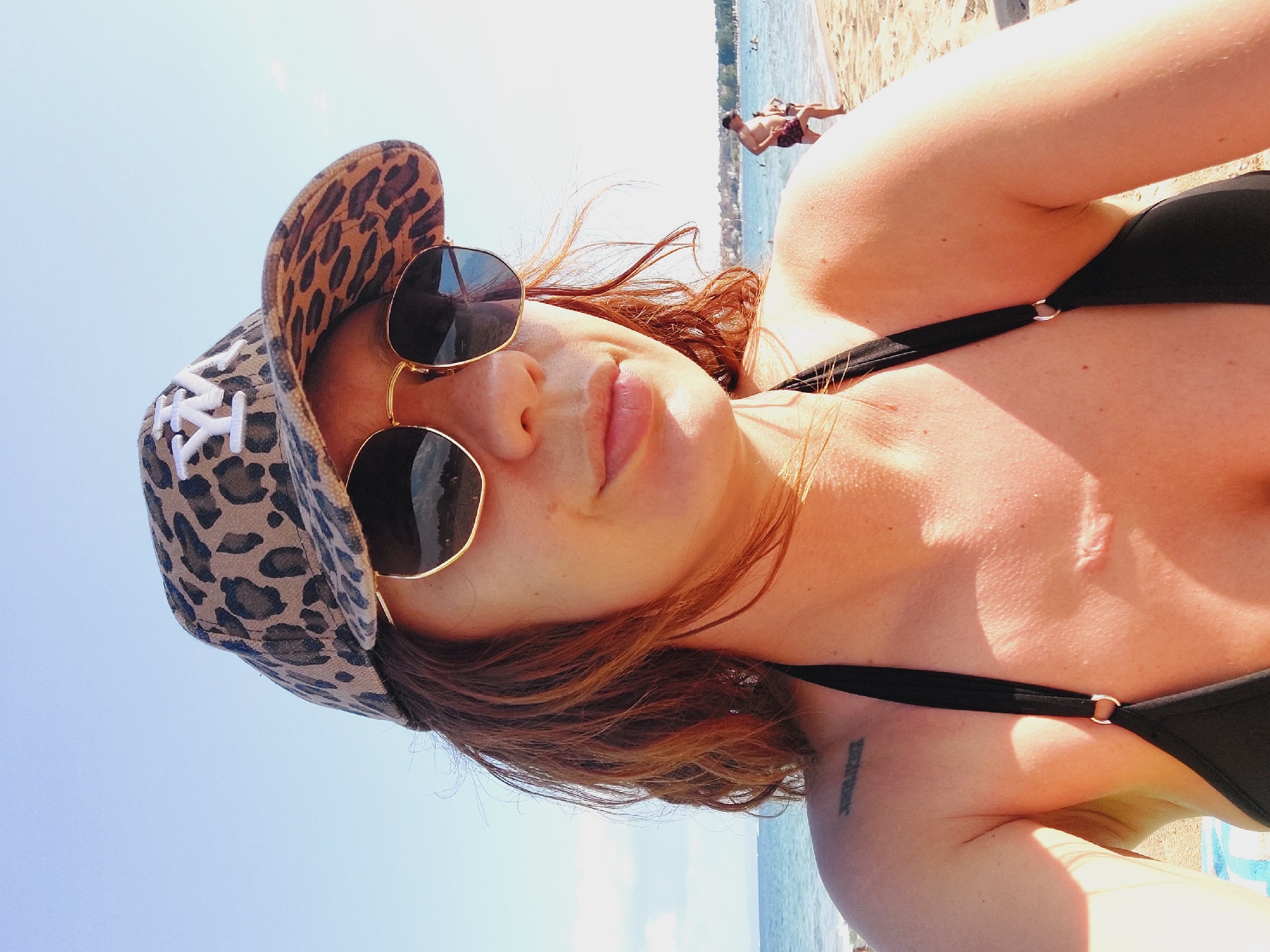 A close-up, chest-up selfie of a woman with auburn hair, wearing a leopard-print baseball cap and sunglasses, poses on a sunny beach in Sozopol, Bulgaria. Her black bikini top is visible. A Roman numeral tattoo is on her left shoulder, and a scar is on her chest. In the background, the sea meets a sandy beach with a few people in the distance. The sky is clear and blue.